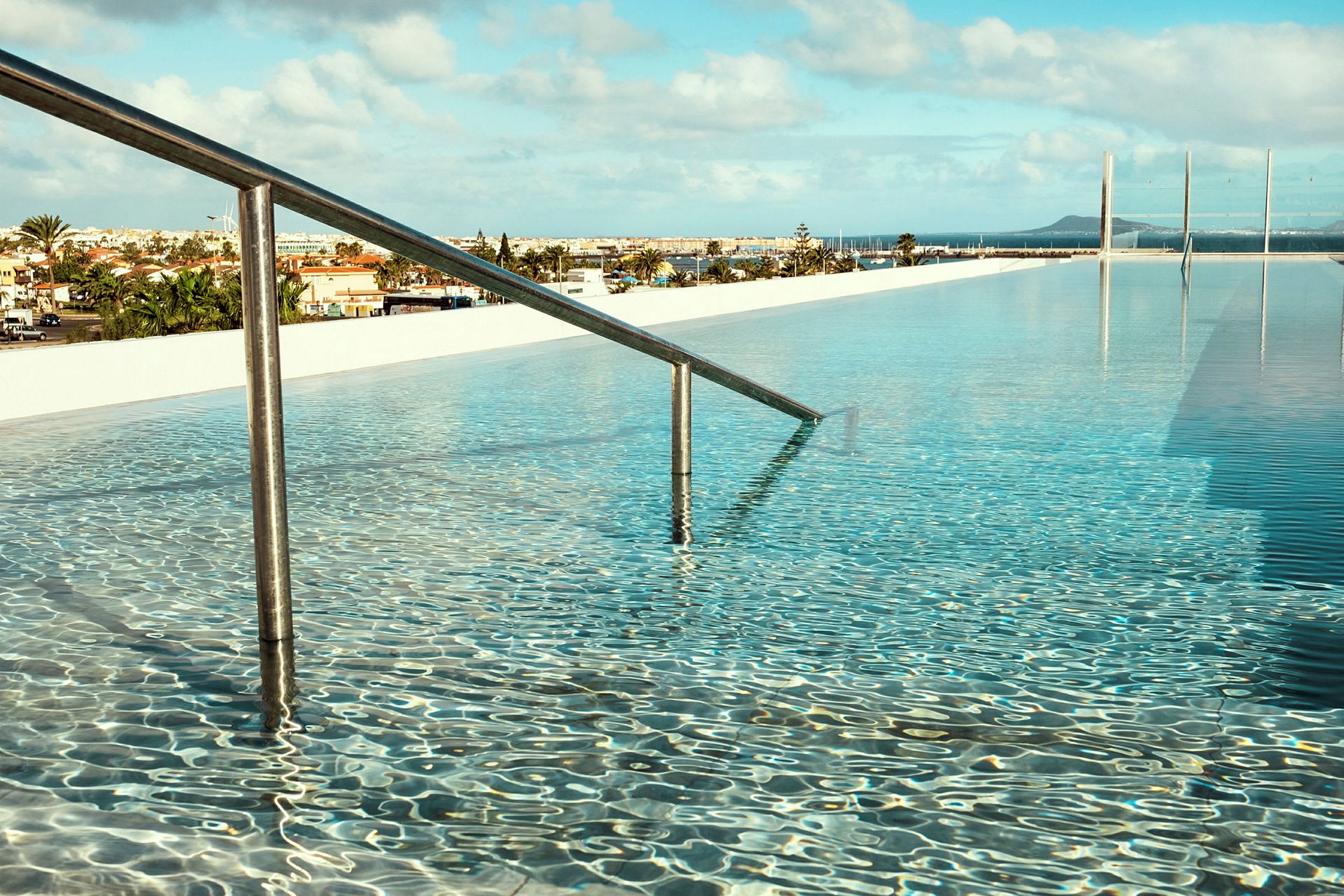 En pool med klart vand og et gelænder, der fører ned i vandet. I baggrunden ses en by og blå himmel med enkelte skyer. (AI-genereret tekst)