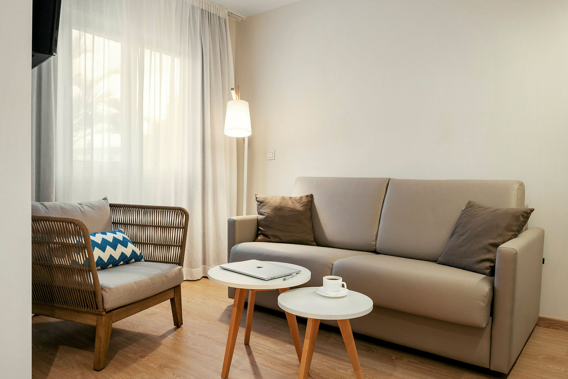 En stue med en beige sofa, en stol med flettet ryglæn, to små hvide borde og en gulvlampe. Et vindue med lyse gardiner ses i baggrunden. (AI-genereret tekst)