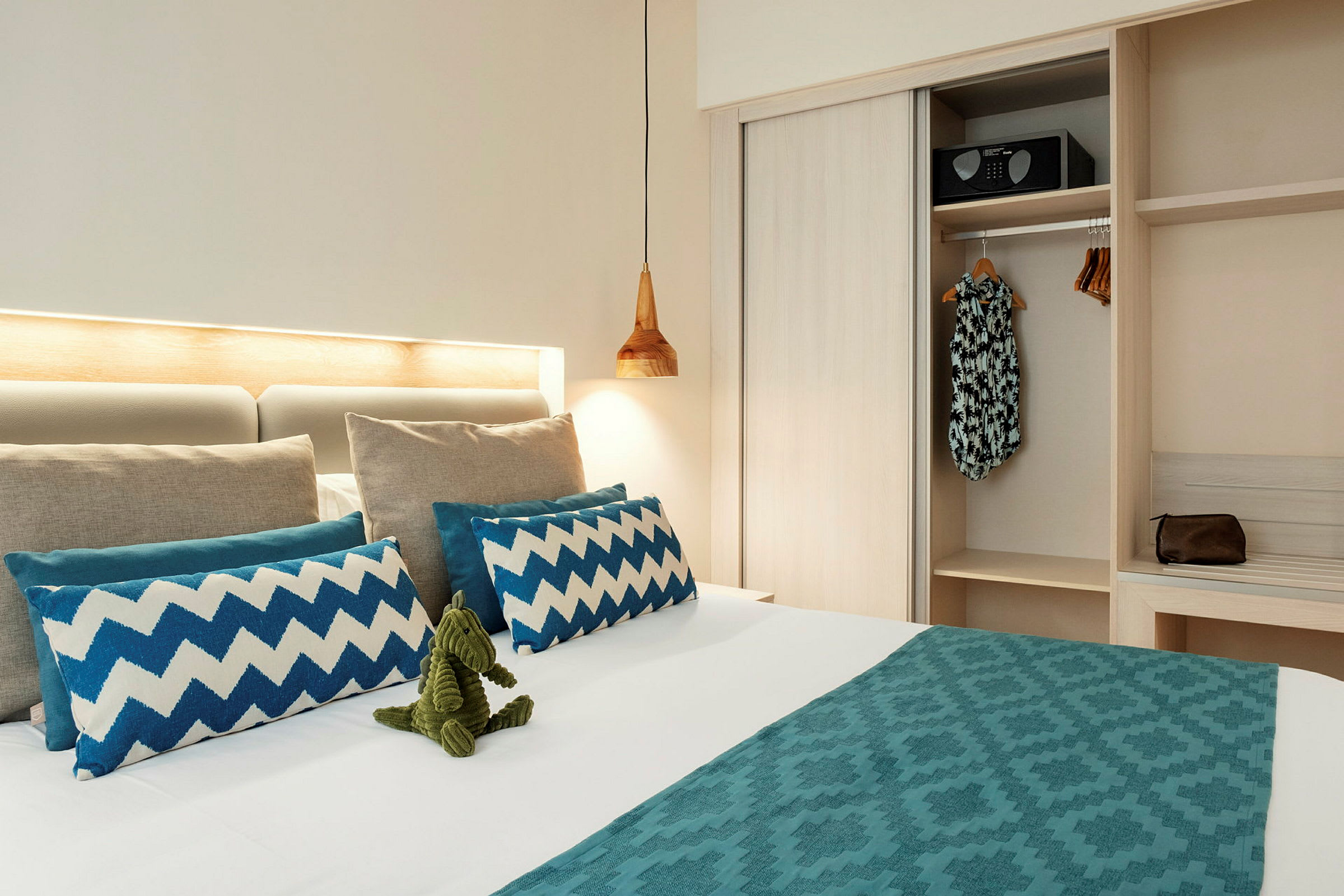 Et hotelværelse med en redt seng, dekoreret med blå puder og en grøn legetøjsfigur. Et skab med tøj og en sikkerhedsboks er synligt ved siden af. (AI-genereret tekst)