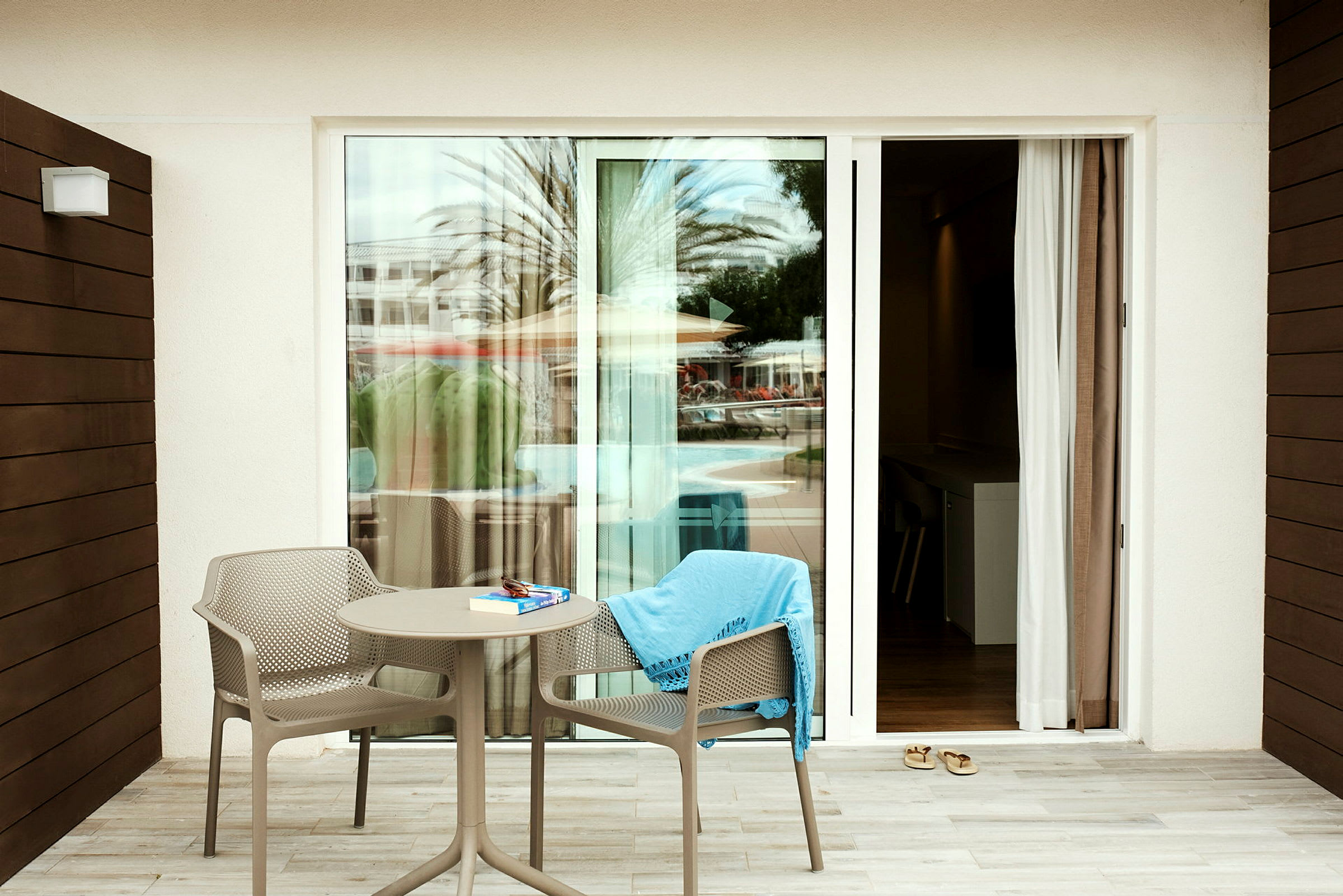 En terrasse med to stole og et bord foran en glasdør, med udsigt til en pool og parasoller. (AI-genereret tekst)