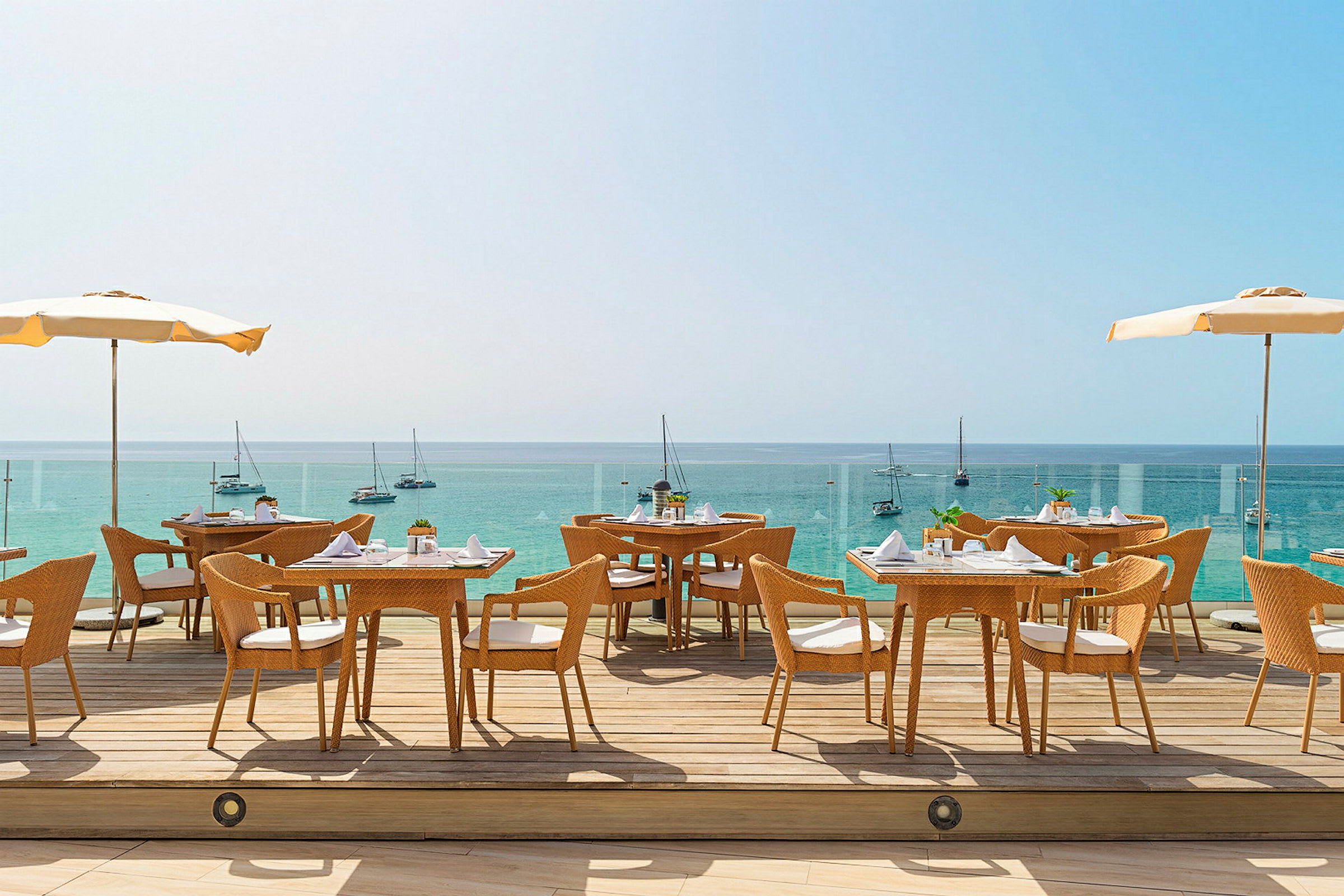 Udendørsrestaurant med udsigt over havet. Flere borde og stole står på en terrasse med parasoller, og sejlbåde kan ses i baggrunden. (AI-genereret tekst)