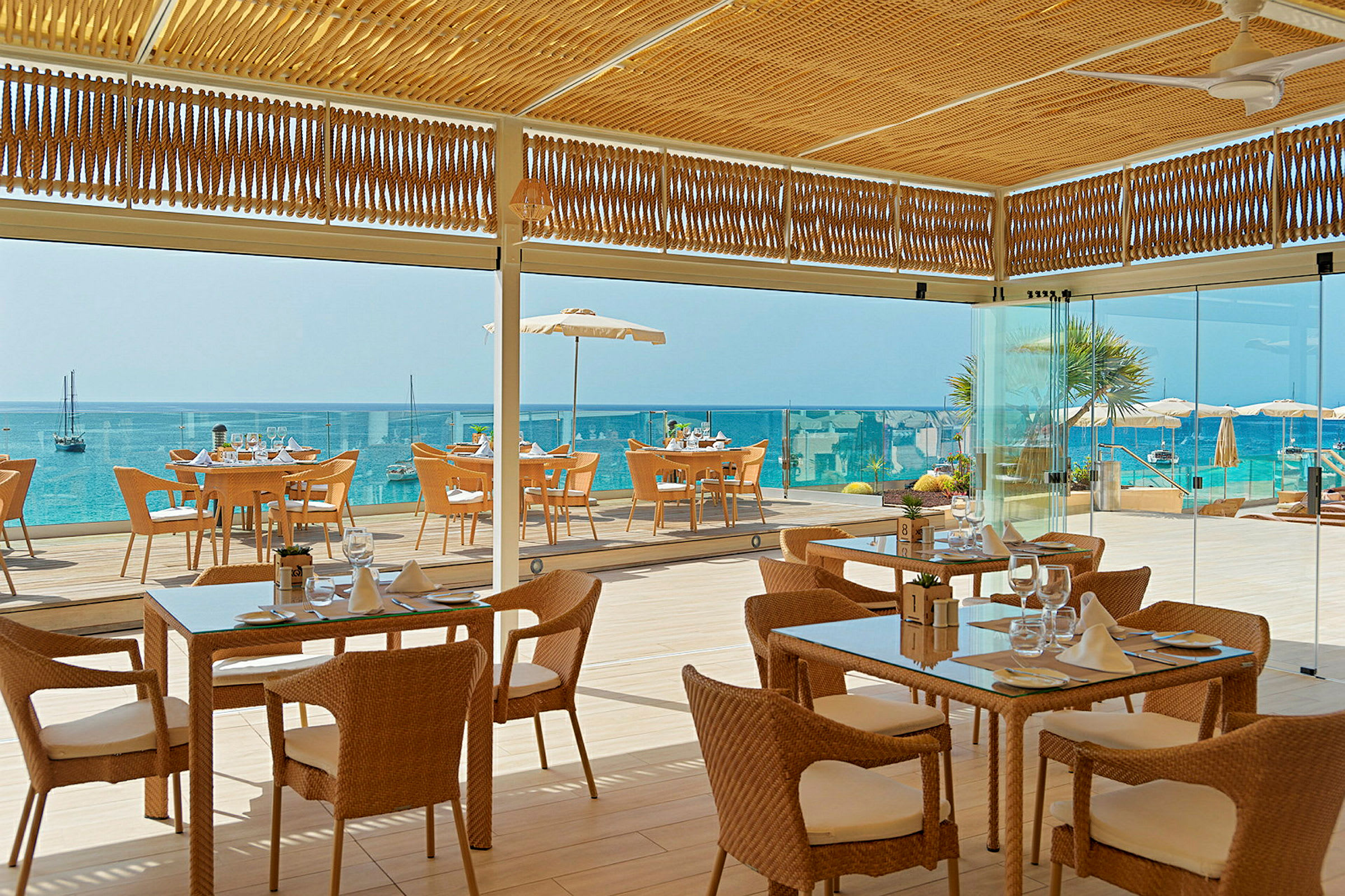 Billede af en restaurant med flettede stole og glasborde, beliggende ved havet med udsigt til en blå vandhorisont. (AI-genereret tekst)