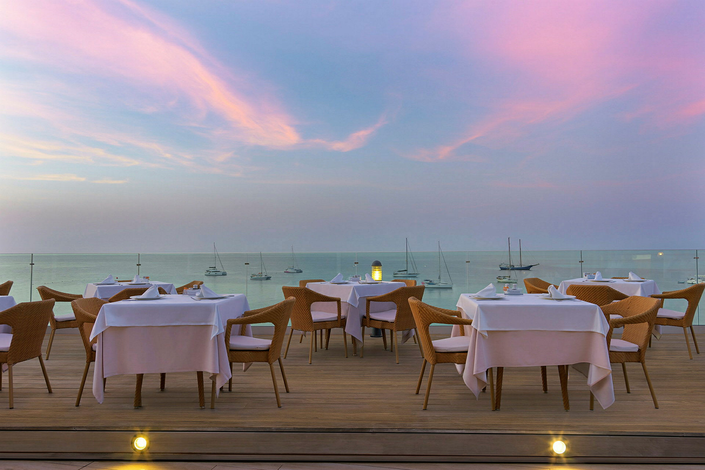 En restaurantterrasse med borde og stole med udsigt over havet under en lyserød og blå aftenhimmel. (AI-genereret tekst)