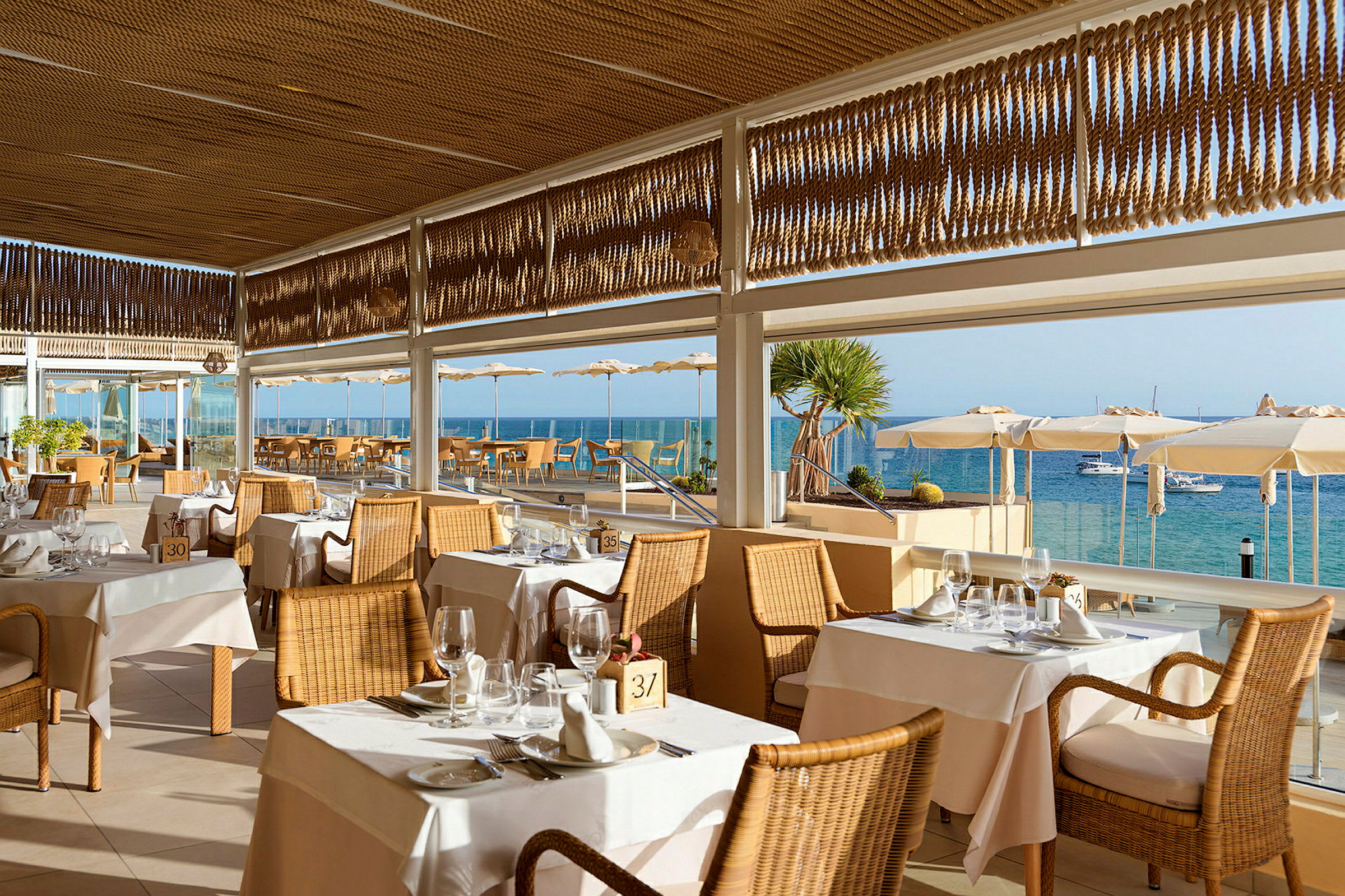 Restaurant med fletstole og opdækkede borde, beliggende ved havet med udsigt over vandet og bådene. (AI-genereret tekst)