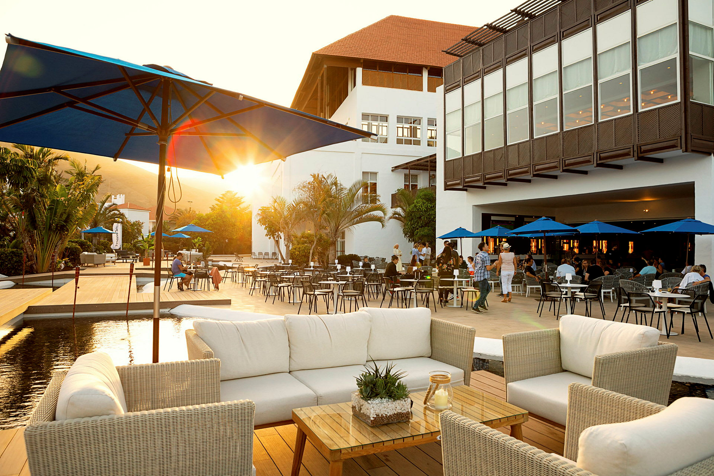 En restaurantterrasse med hvide rottingmøbler og parasoller, omgivet af et palmklædt område og en stor hvid bygning i baggrunden. (AI-genereret tekst)