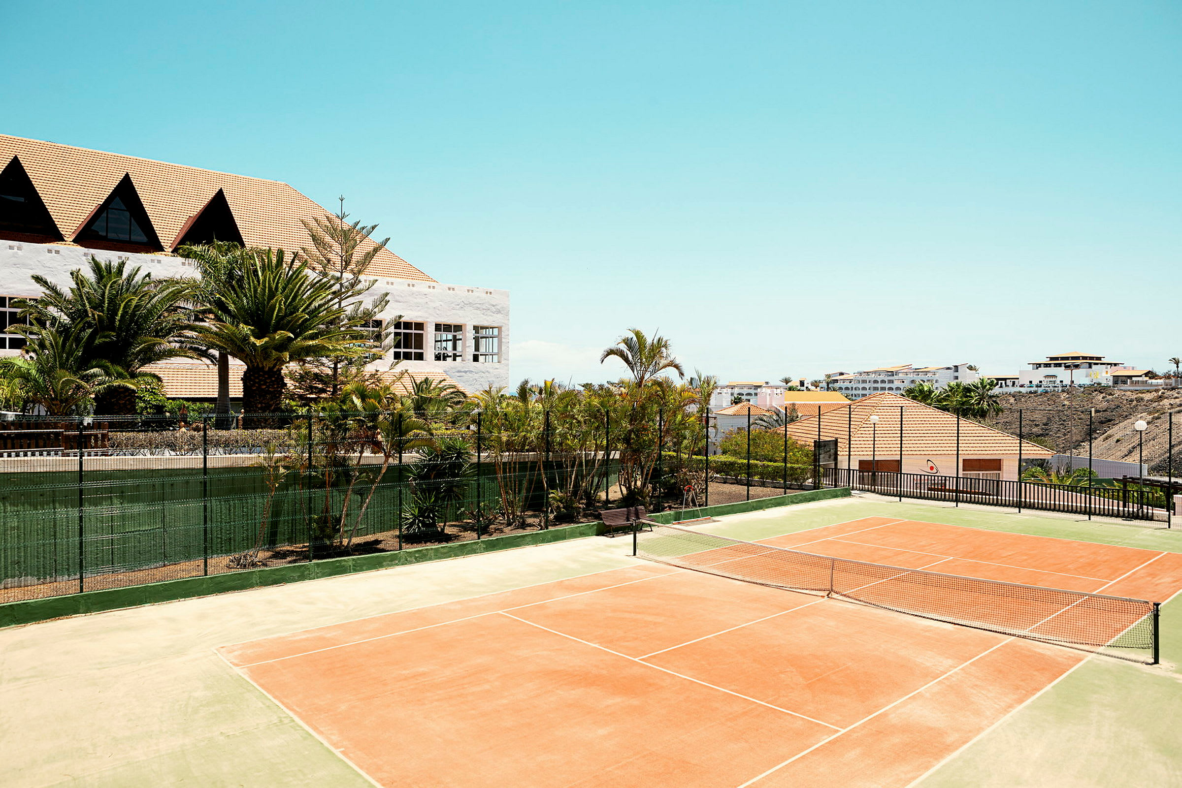 En tennisbane i solen med omkringliggende beplantning og bygninger i baggrunden. (AI-genereret tekst)