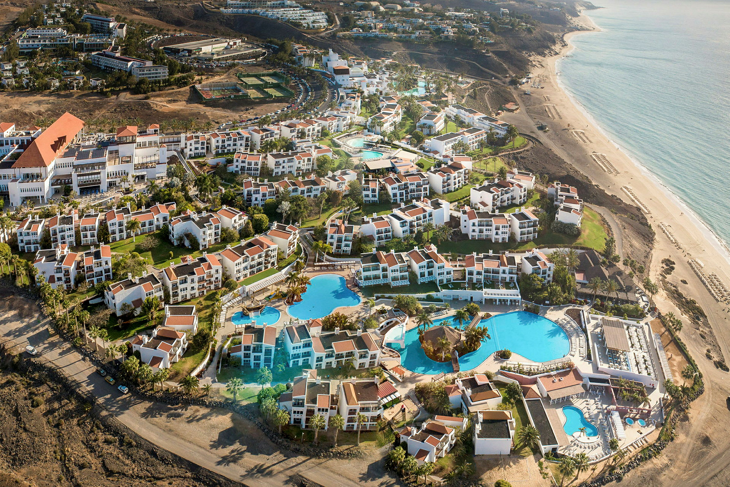 Luftfoto af et feriested med hvide bygninger og røde tage, samt store swimmingpools og haver nær stranden. (AI-genereret tekst)