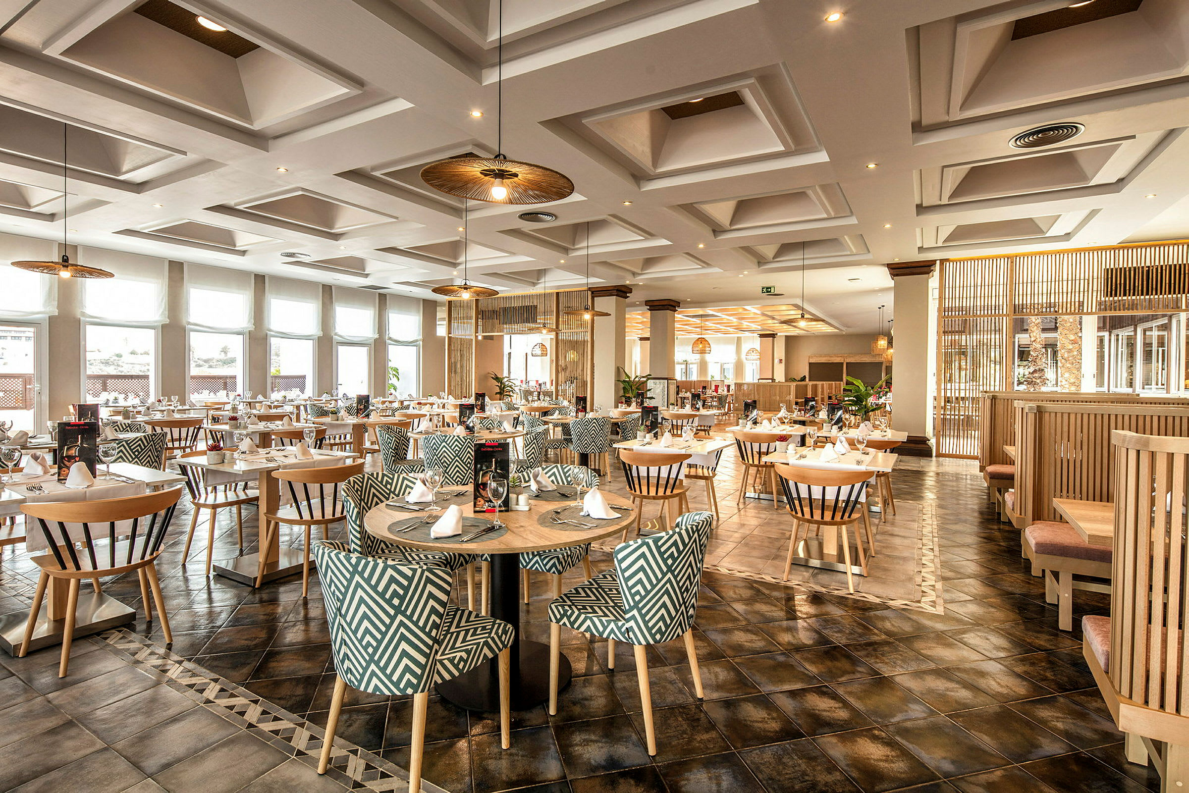 Et elegant restaurantområde med runde borde og mønstrede stole, belyst af loftslamper og naturligt lys gennem store vinduer. (AI-genereret tekst)
