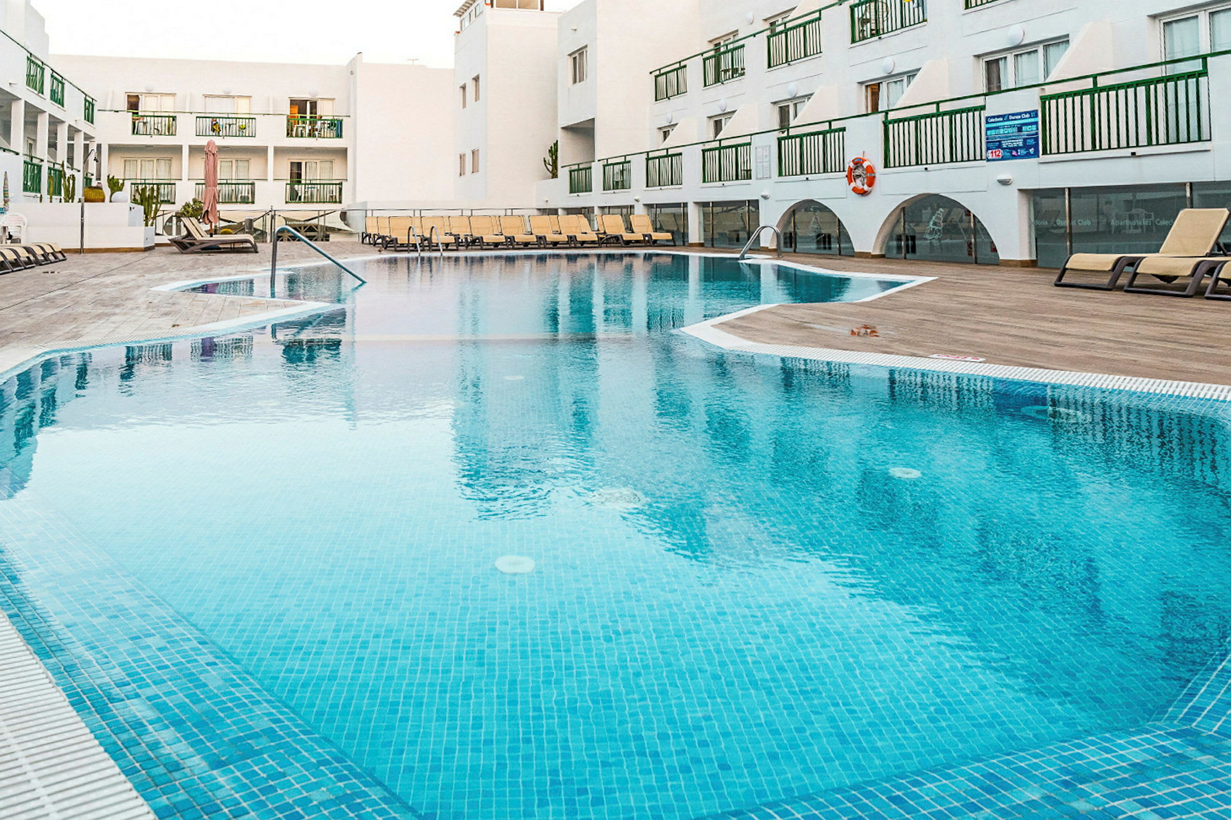 Udendørs swimmingpool på et feriested med liggestole og hvide bygninger i baggrunden. (AI-genereret tekst)