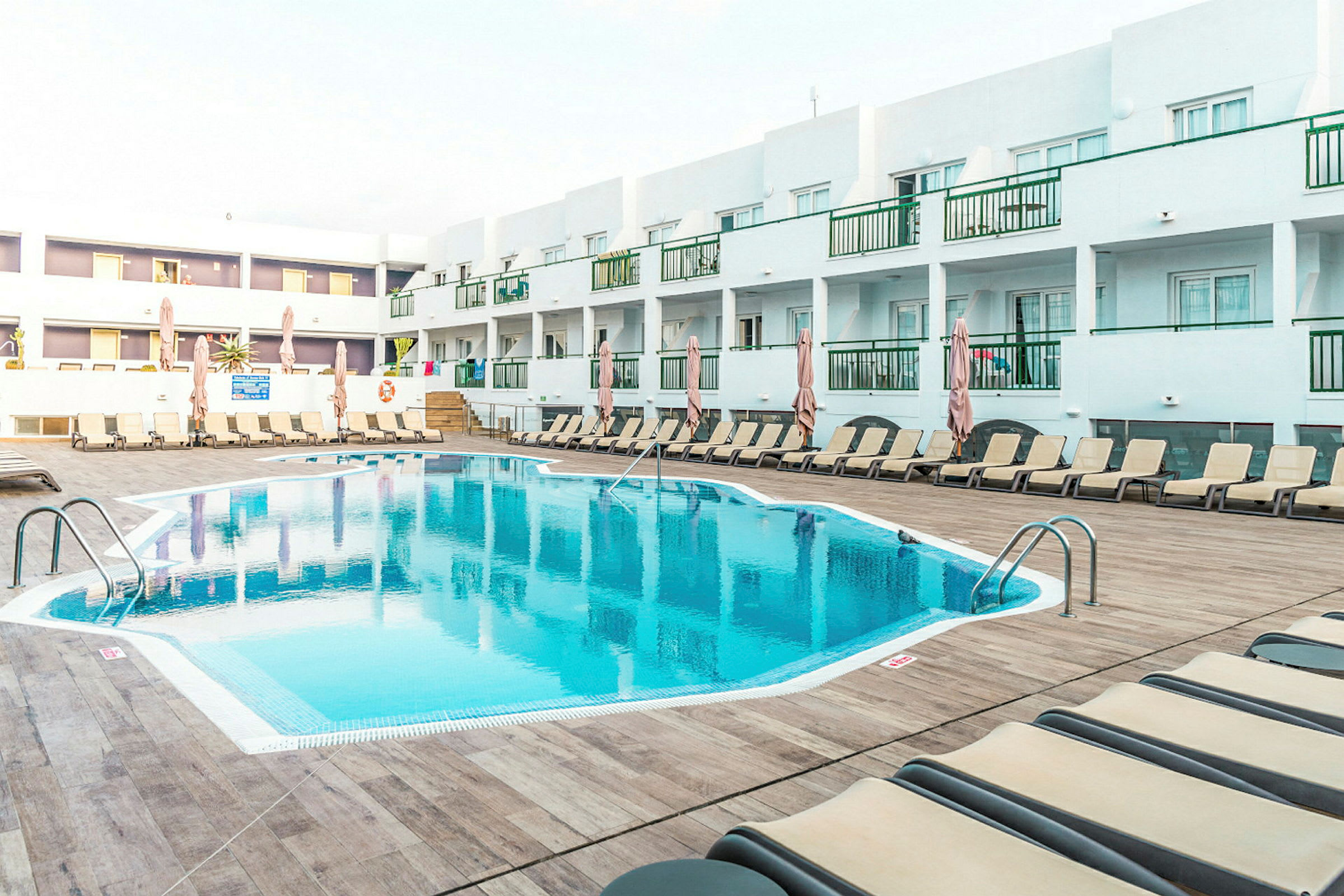 Udendørs pool ved et hotel omgivet af liggestole og parasoller, med hvide bygninger i baggrunden. (AI-genereret tekst)
