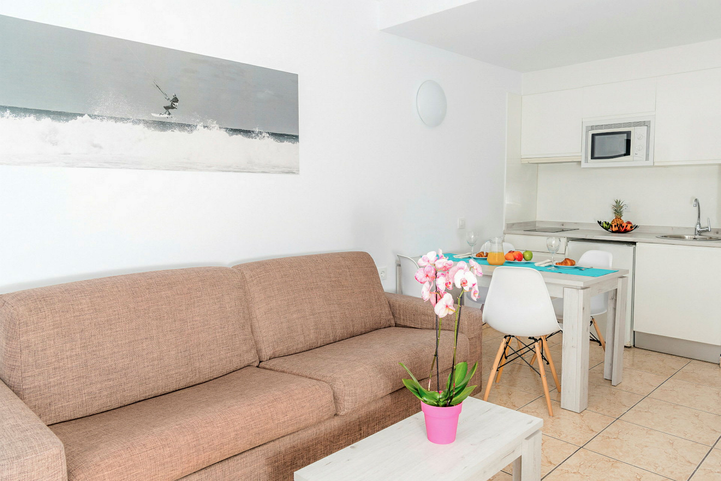 Et moderne rum med en brun sofa, et sofabord med en lyserød potteplante, et køkken med hvide skabe og et spiseområde med et dækket bord. (AI-genereret tekst)