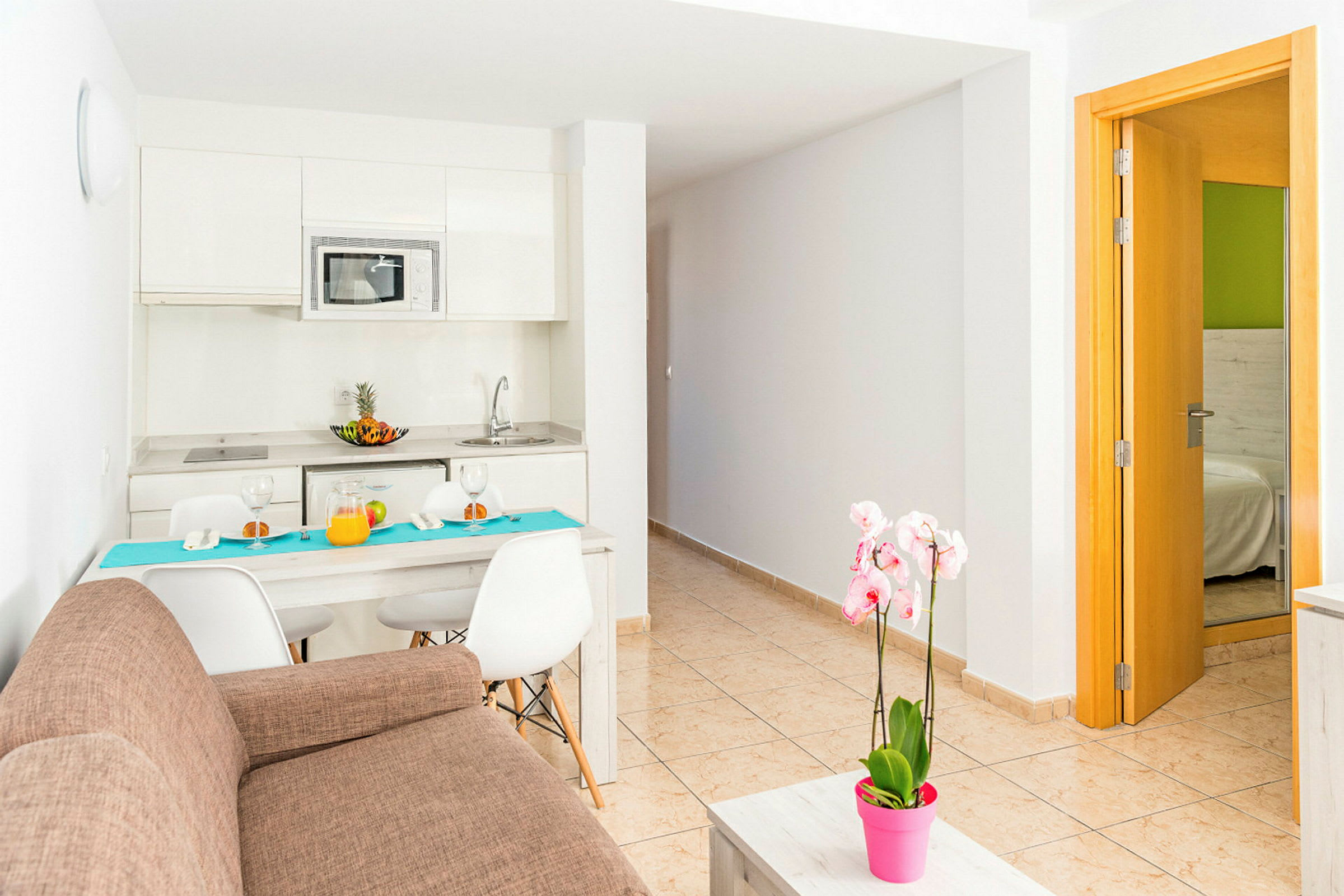 Et moderne køkken med spisebord og stole, en brun sofa og en pink blomsterpotte på et sofabord. Indgang til soveværelse er synlig til højre. (AI-genereret tekst)