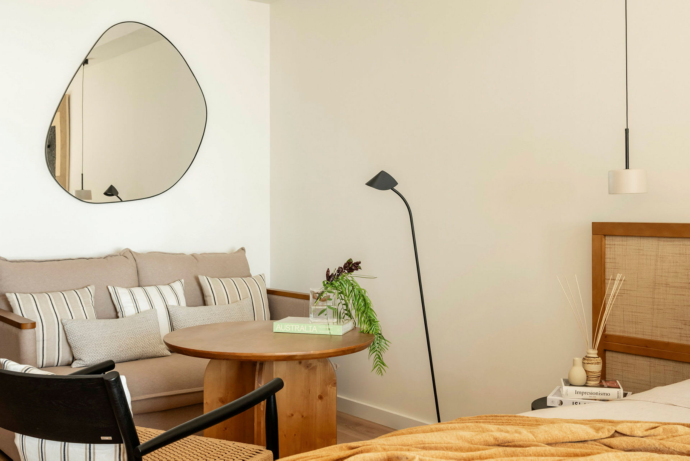 Et rum med moderne interiør. En sofa med puder, et rundt træbord, en gulvlampe og et spejl på væggen. Til højre ses en seng og et natbord. (AI-genereret tekst)