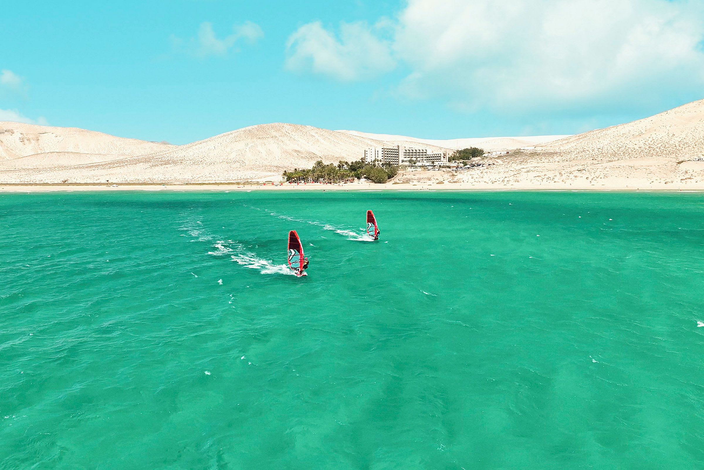 To windsurfere på blågrønt vand med et sandet landskab og en bygning i baggrunden. (AI-genereret tekst)