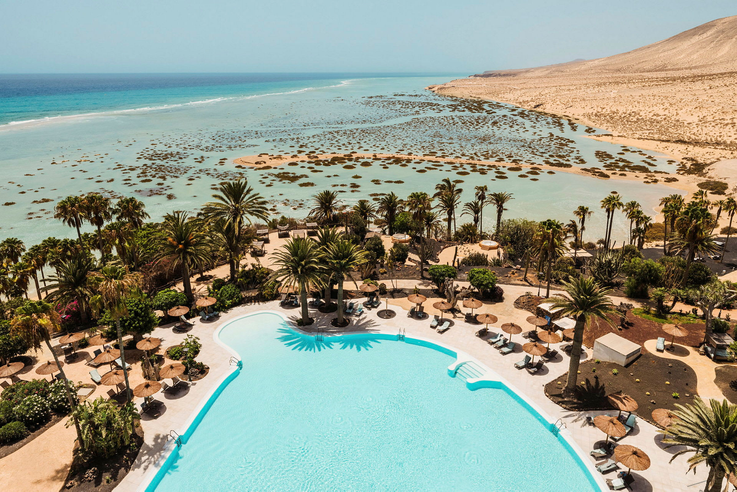 En pool omgivet af liggestole og parasoller med palmer i baggrunden samt udsigt over en lavvandet turkisblå strand og hav. (AI-genereret tekst)