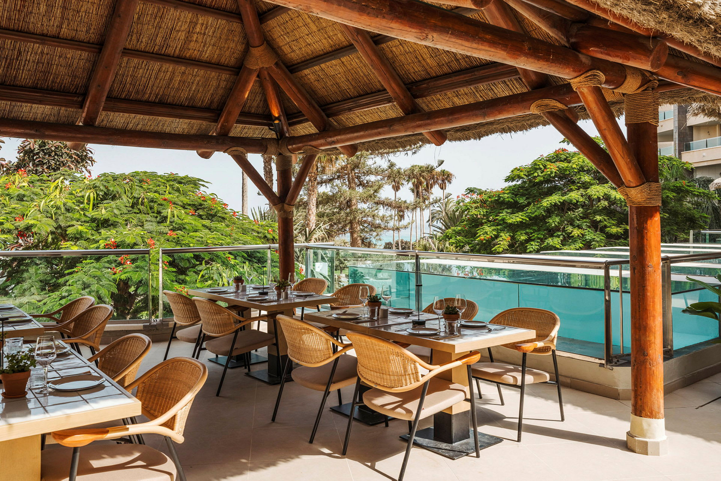 Restaurant med borde og stole under trætag med udsigt til grønne områder og pool. (AI-genereret tekst)