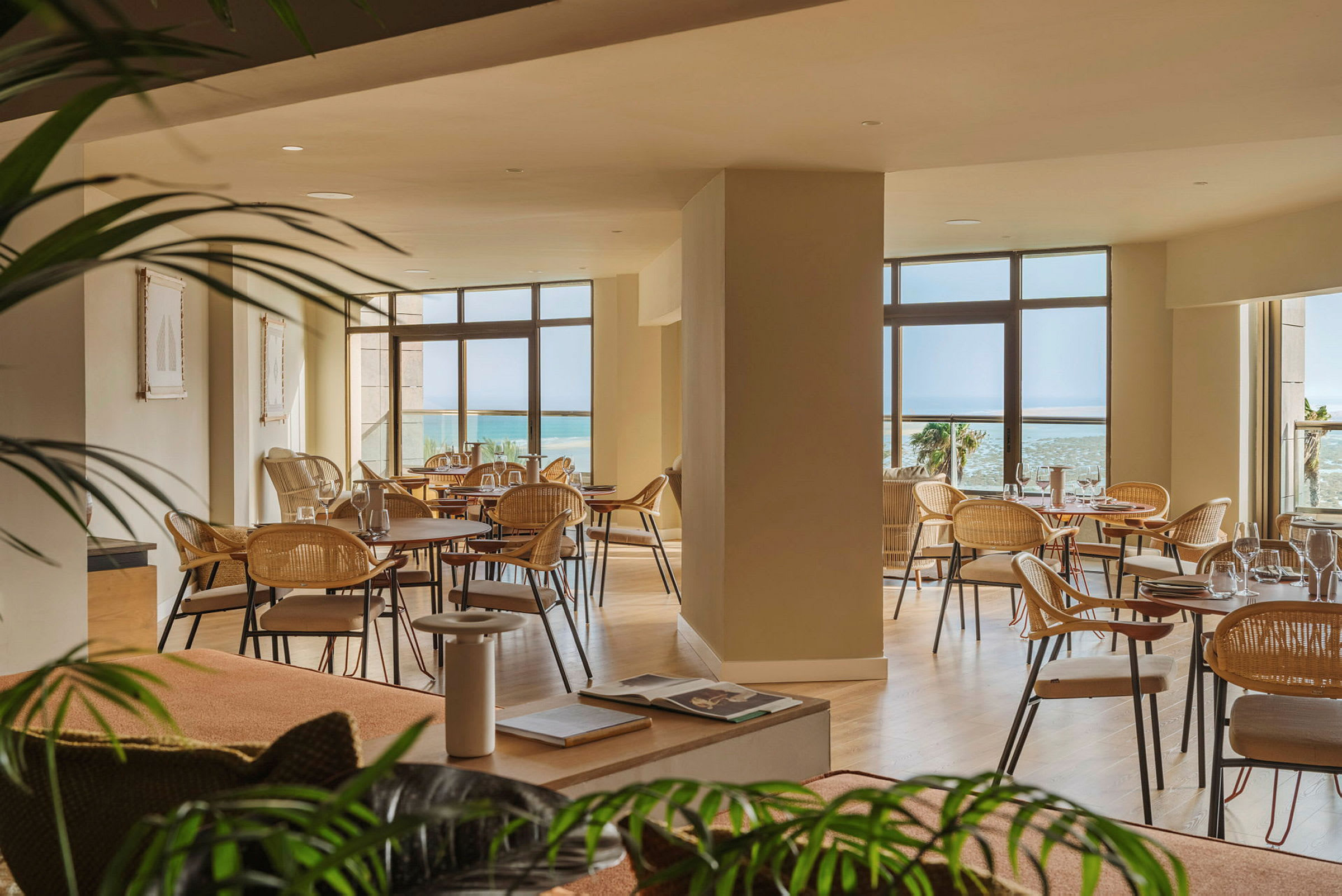 En rummelig restaurant med store vinduer, der giver udsigt til havet. Der er flere borde med stole, og indretningen er lys og elegant. (AI-genereret tekst)
