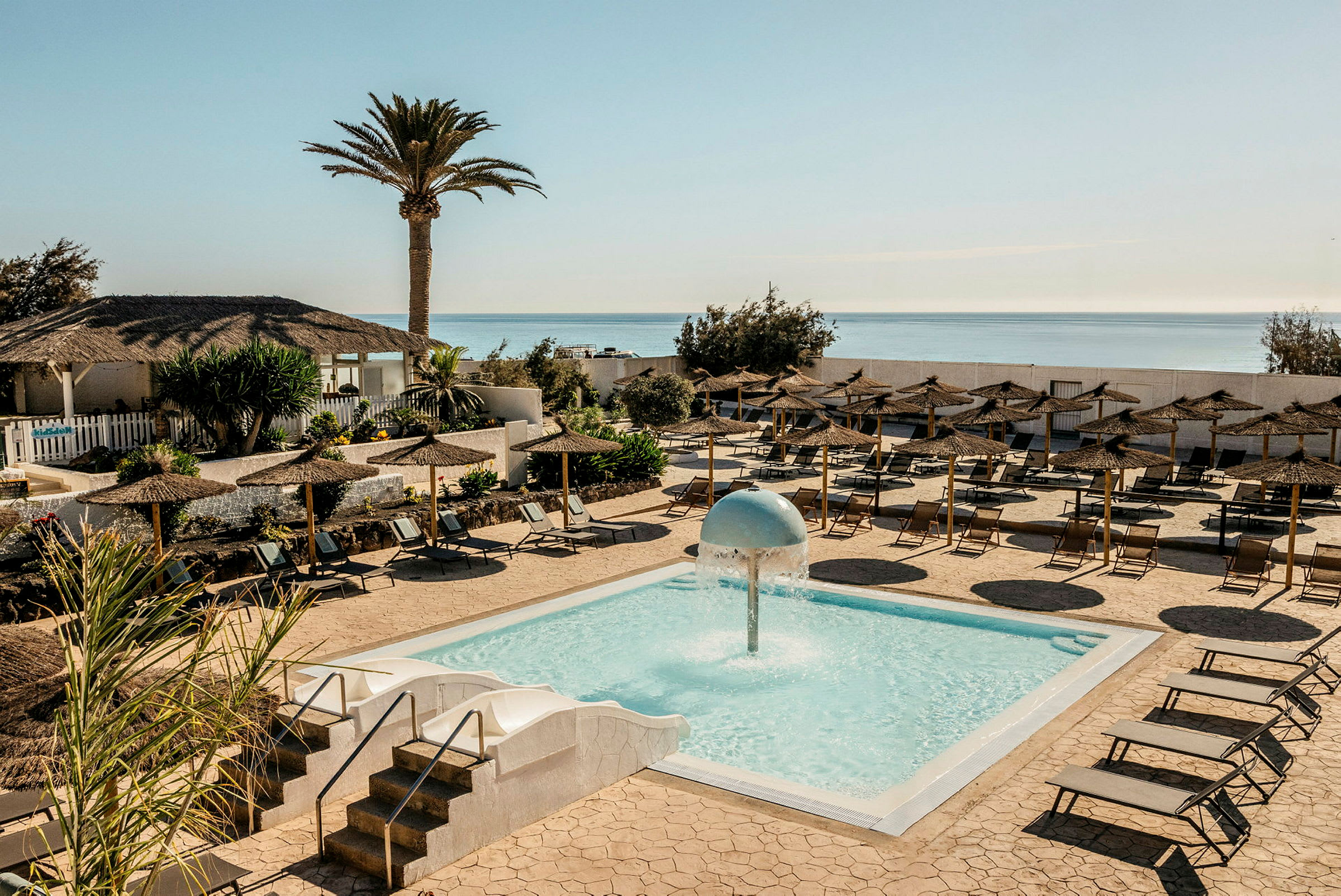 En udendørs pool med en vandfontæne i midten, omgivet af liggestole og parasoller. I baggrunden ses en palme, en bygning med stråtag og havet. (AI-genereret tekst)