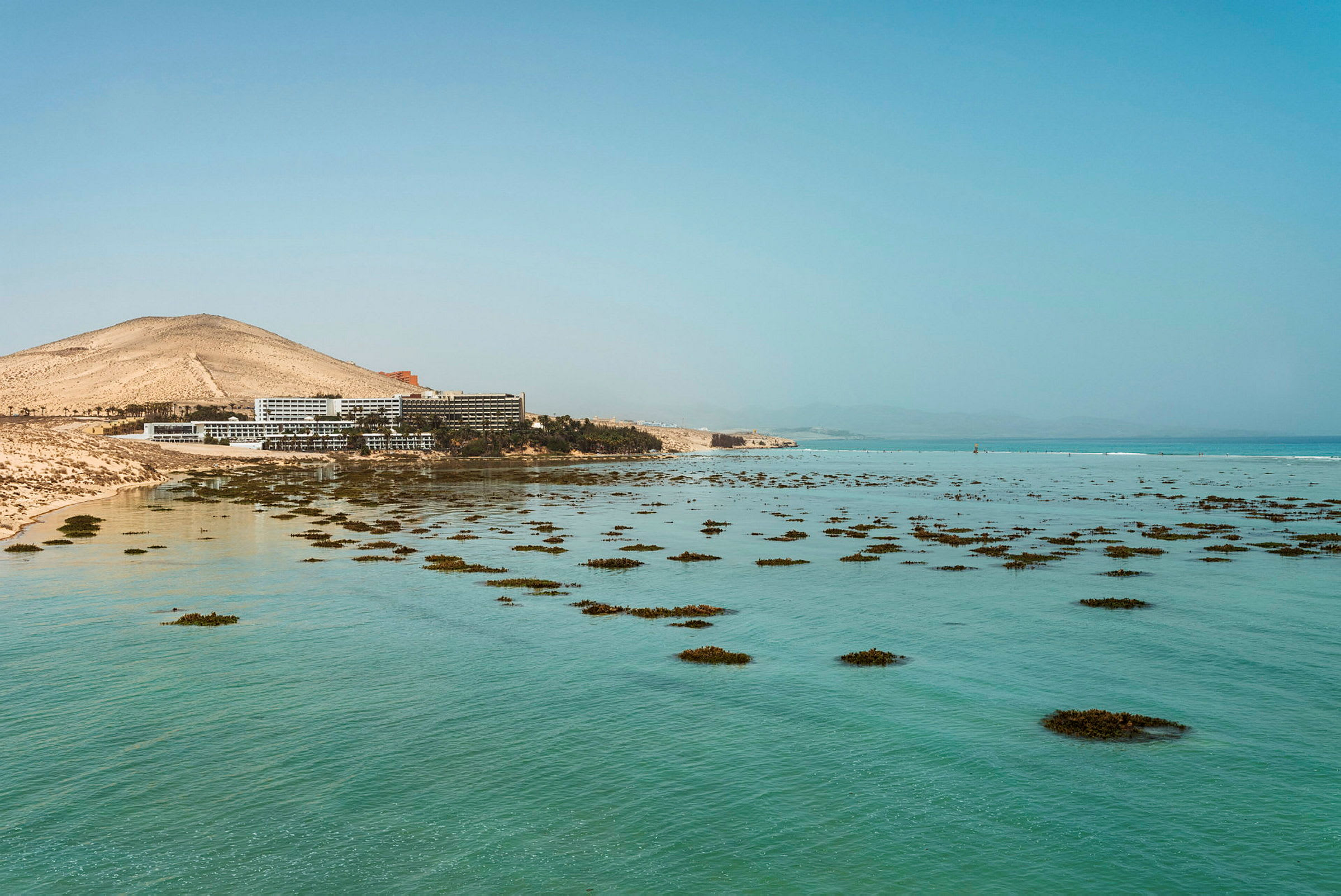 Hotelbygninger nær en strand med klart blåt vand og spredte klynger af vegetation. (AI-genereret tekst)