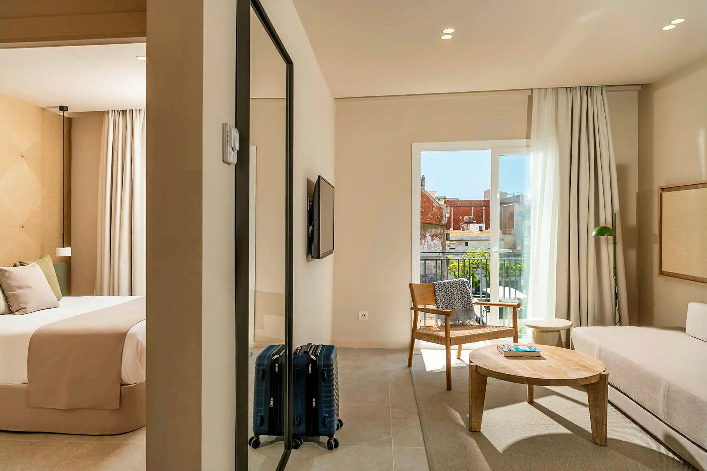 Et hotelværelse med et soveværelse og opholdsstue. Soveværelset har en seng med beige sengetøj, og opholdsstuen har en sofa, et træbord og en lænestol. Der er en balkon med udsigt. (AI-genereret tekst)