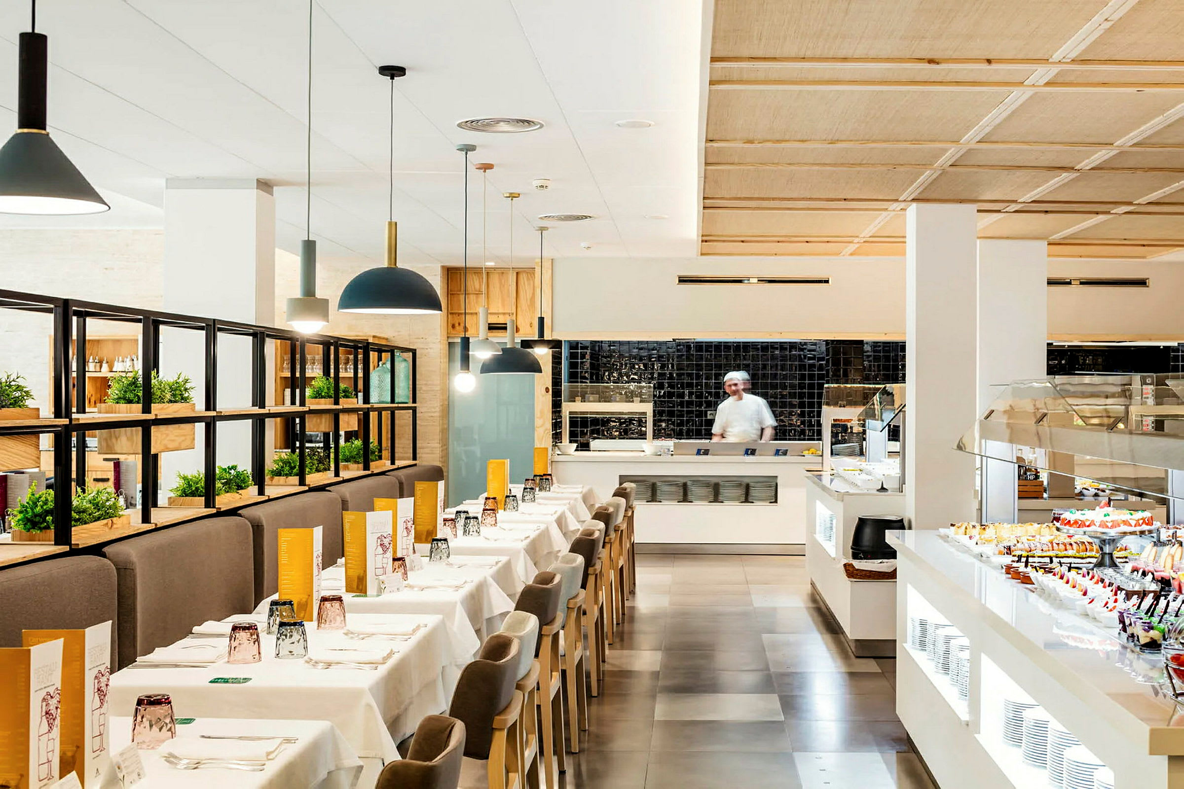 En lys restaurant med dækkede borde, en serveringsbuffet og en kok i køkkenet. (AI-genereret tekst)