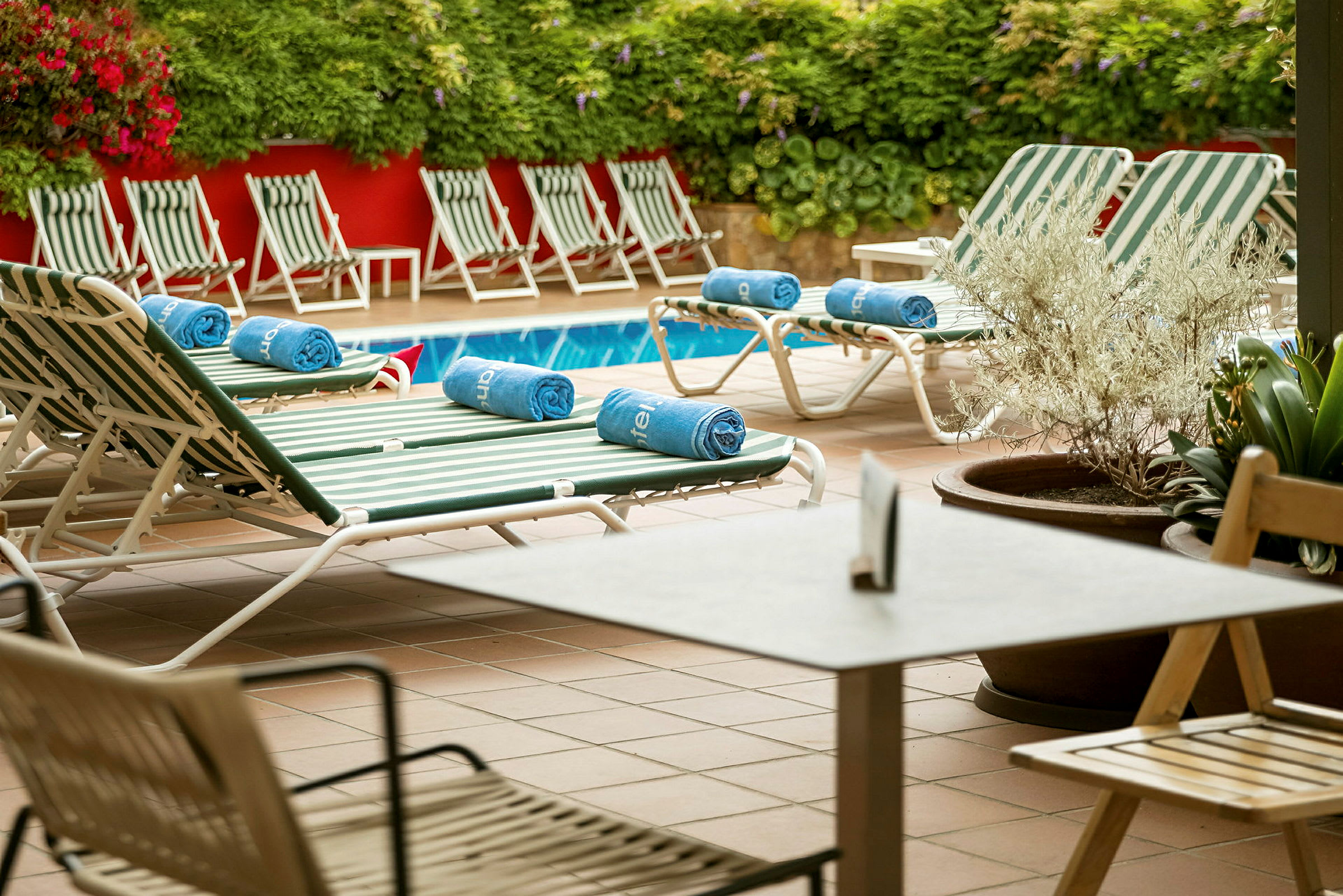 Liggestole og borde ved en pool med grøn beplantning og røde vægge i baggrunden. (AI-genereret tekst)