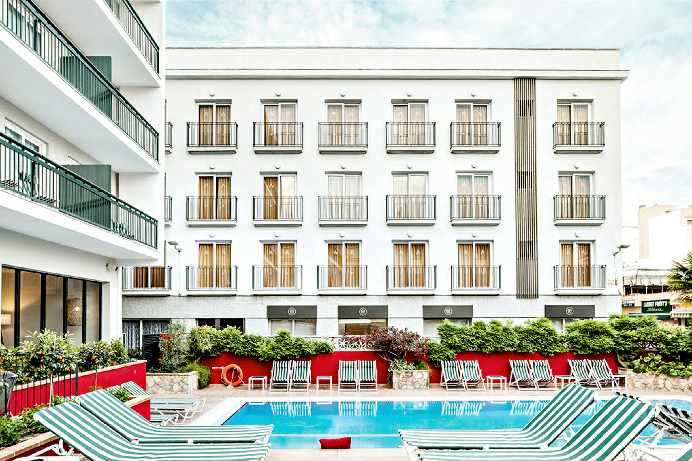 Billede af en hotelbygning med hvid facade og flere altaner. Foran bygningen er en udendørs pool med stribede liggestole placeret omkring. (AI-genereret tekst)