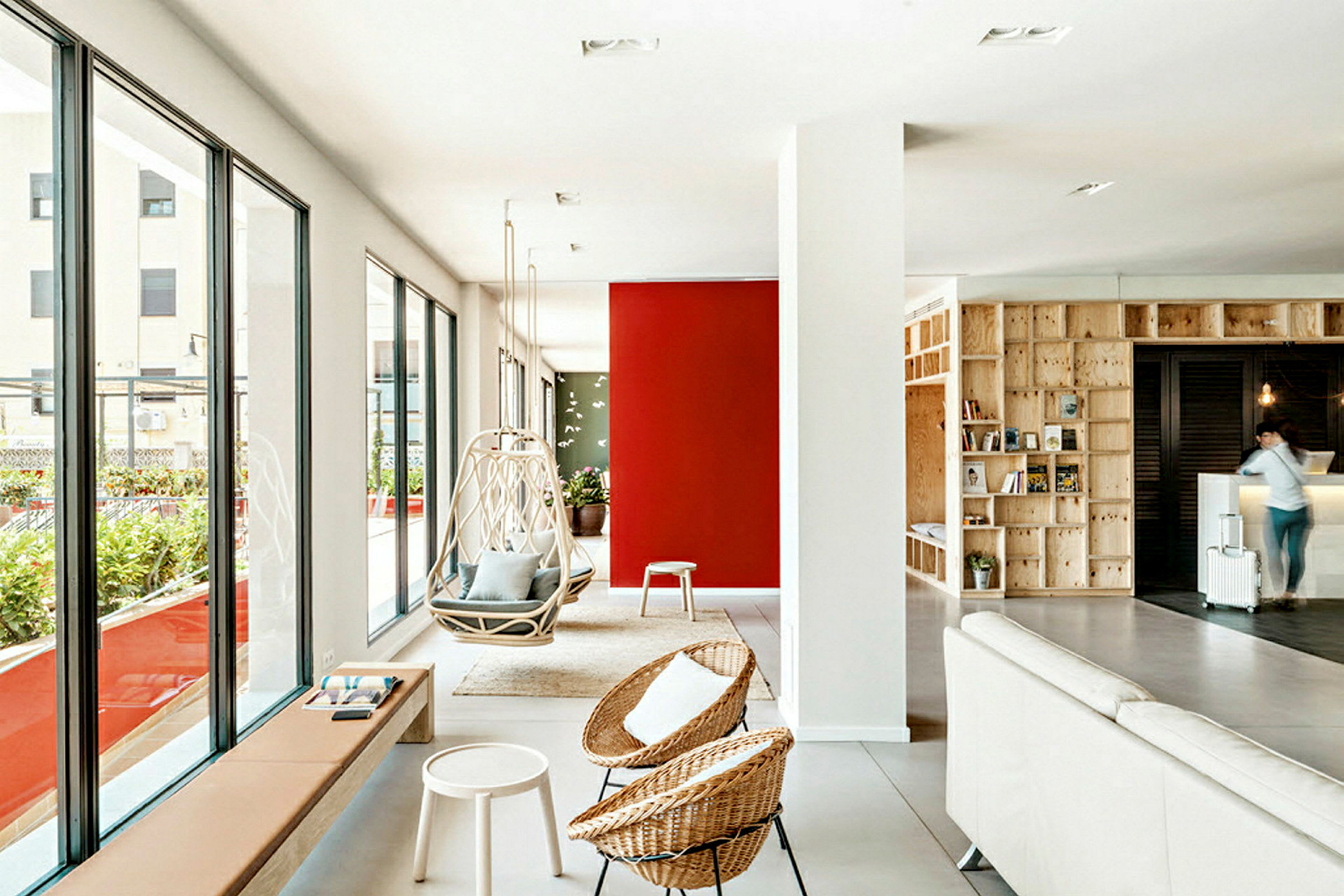 Et moderne og lyst indendørs rum med store vinduer, der vender ud mod en terrasse. Rødt og mønstret på væggen giver rummet farve. Hængestole står sammen med andre designmøbler og detaljer. (AI-genereret tekst)