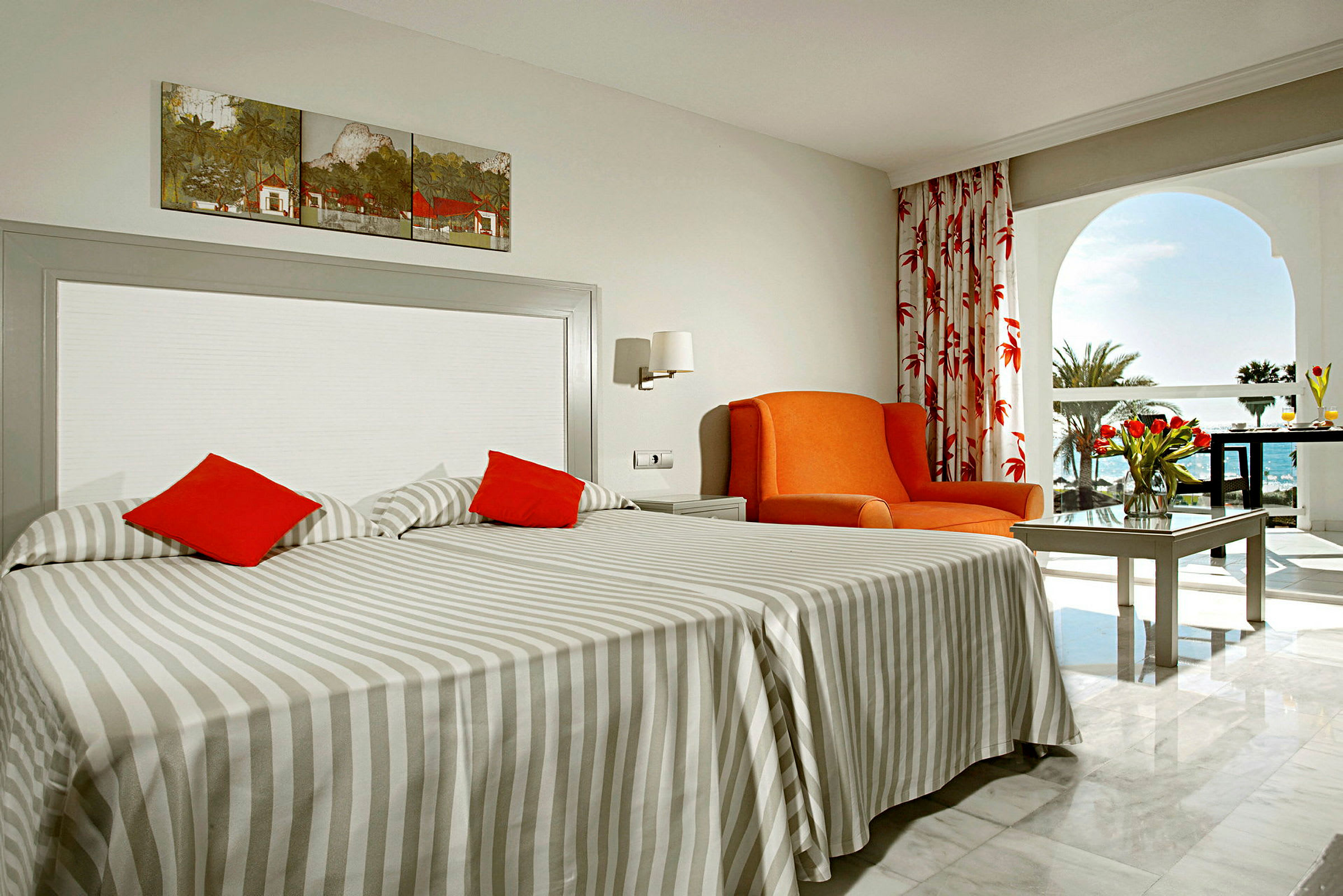 Et hotelværelse med to enkeltsenge, en orange lænestol og et bord med en buket tulipaner. Bag rummet ses en balkon med udsigt over havet. (AI-genereret tekst)