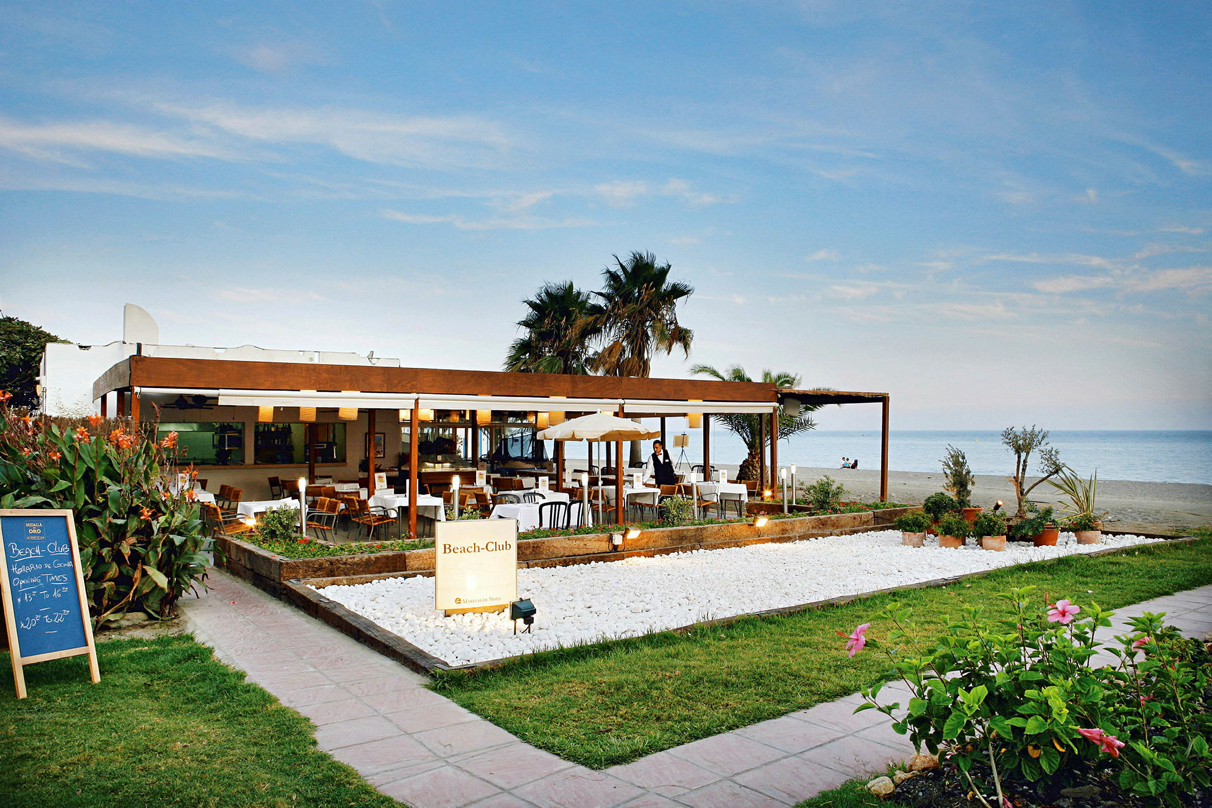 Restaurant med udsigt over stranden og havet, omgivet af planter og med en udendørs terrasse. (AI-genereret tekst)