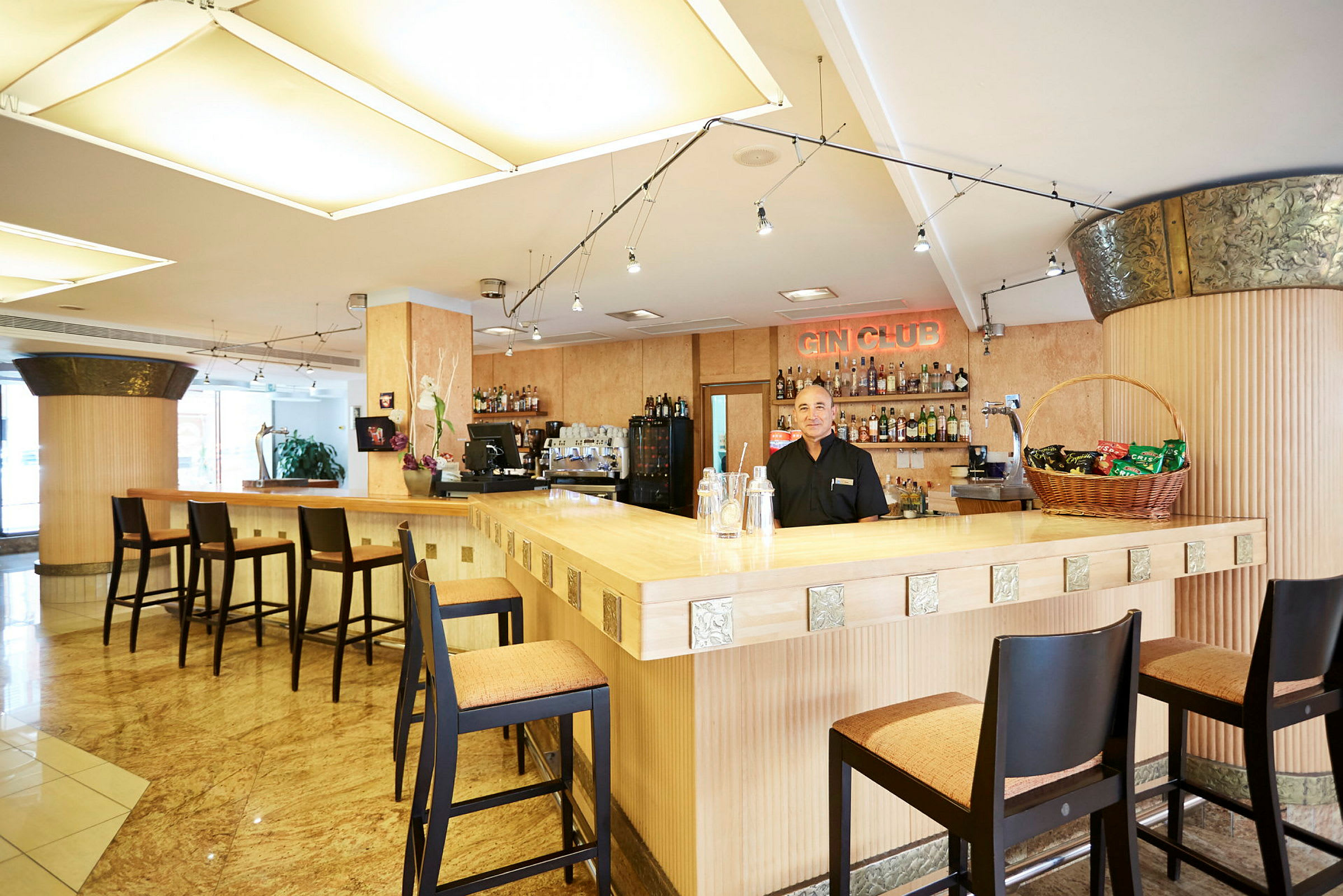 En lys barindretning med høje stole omkring en træbar. På bagvæggen ses flere flasker og et skilt med teksten "Gin Club". (AI-genereret tekst)