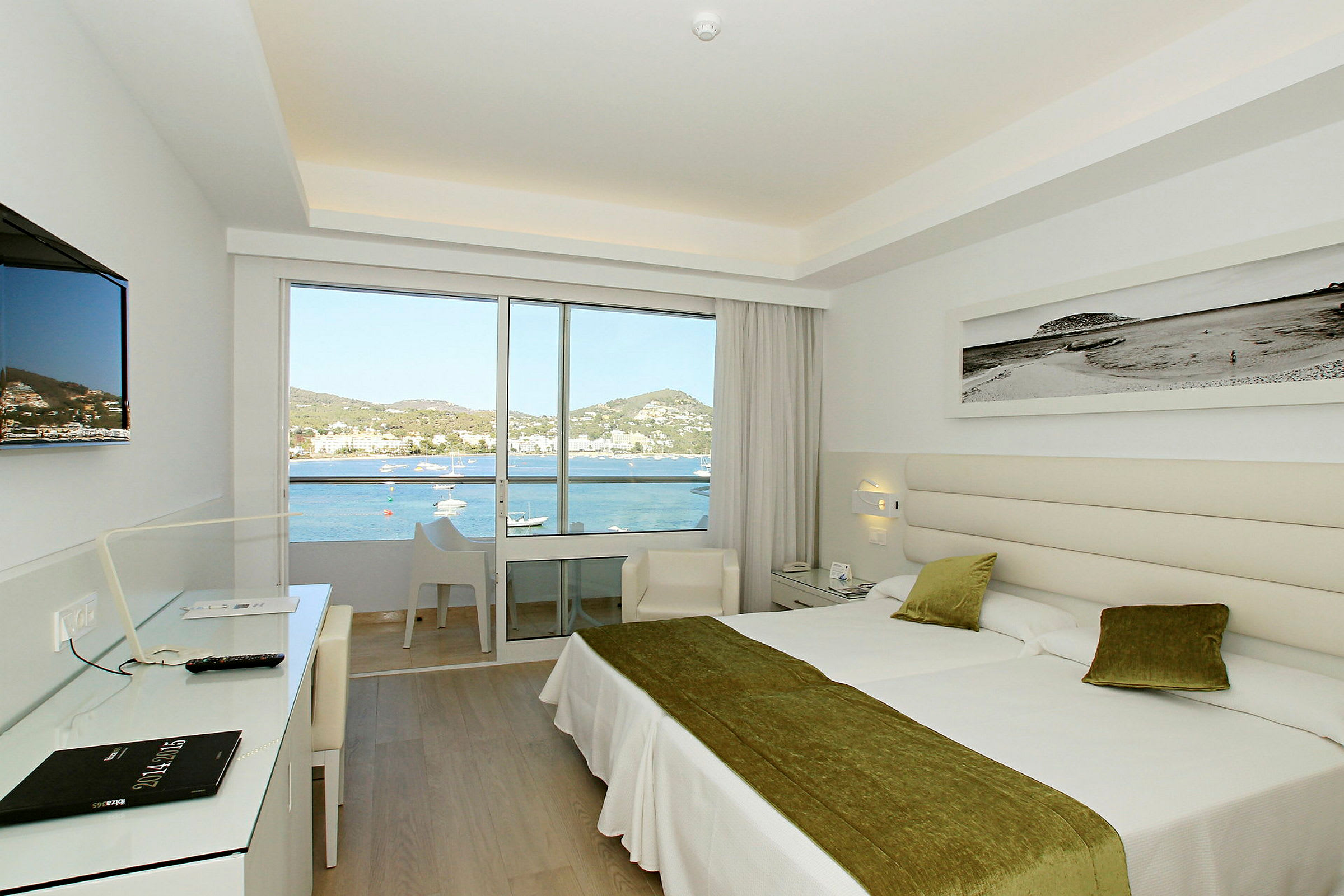 Et lyst og moderne hotelværelse med to senge og store glasdøre, der fører ud til en balkon. Udenfor er der udsigt over havet og det omkringliggende landskab. (AI-genereret tekst)
