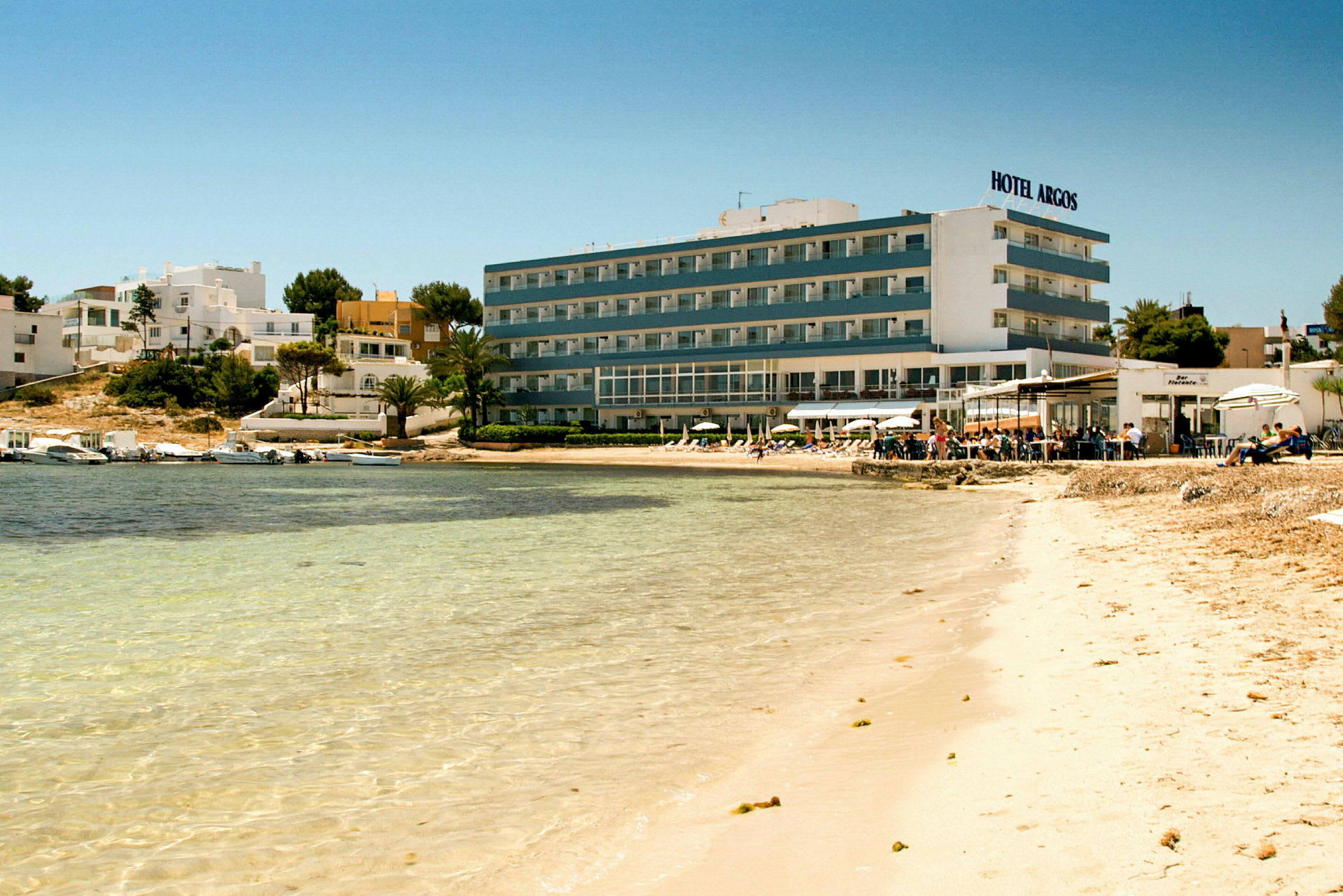 Strand med klart vand foran et hotel og bygninger. (AI-genereret tekst)