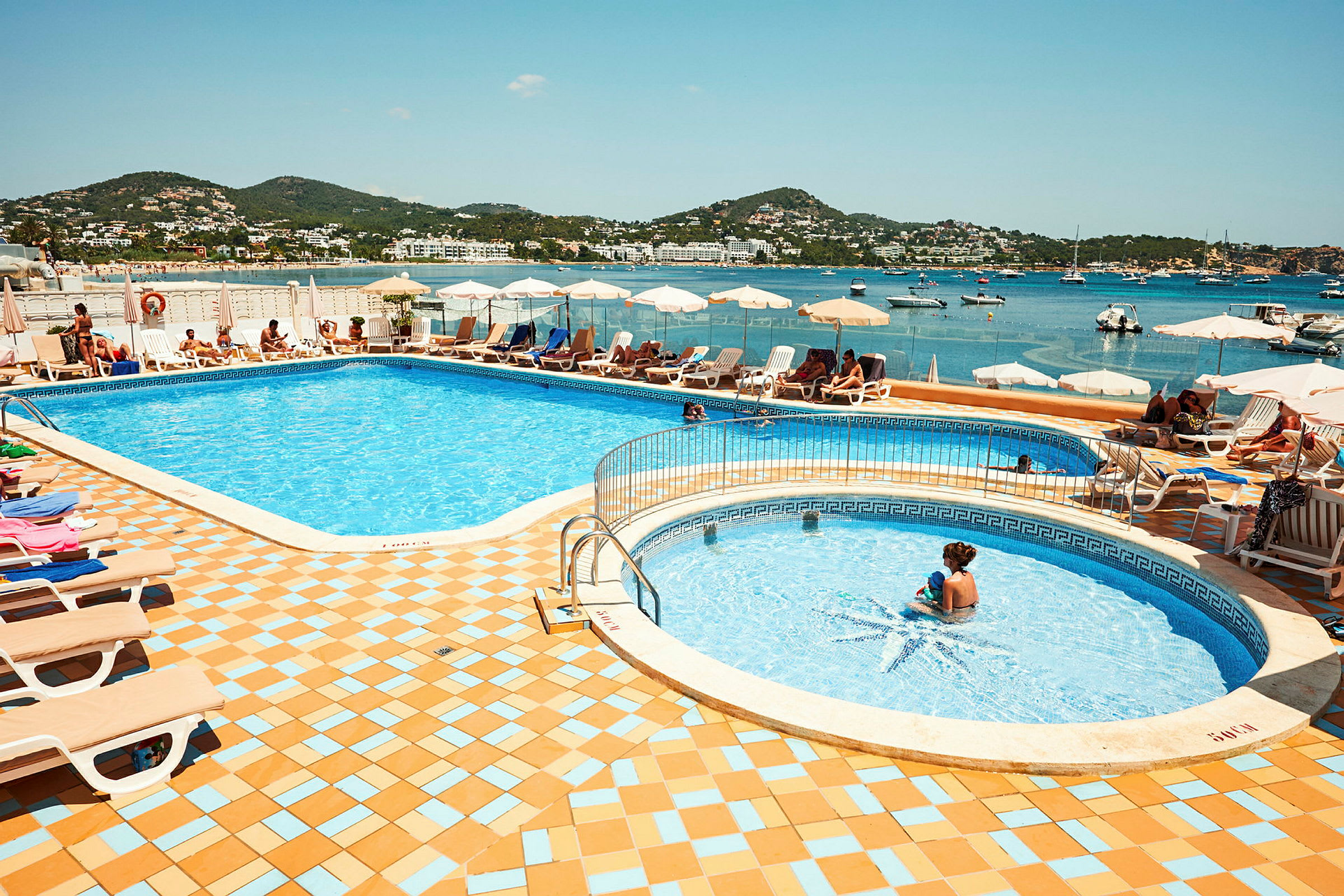 En udendørs swimmingpool med liggestole og parasoller i nærheden, med hav og landskab i baggrunden. (AI-genereret tekst)