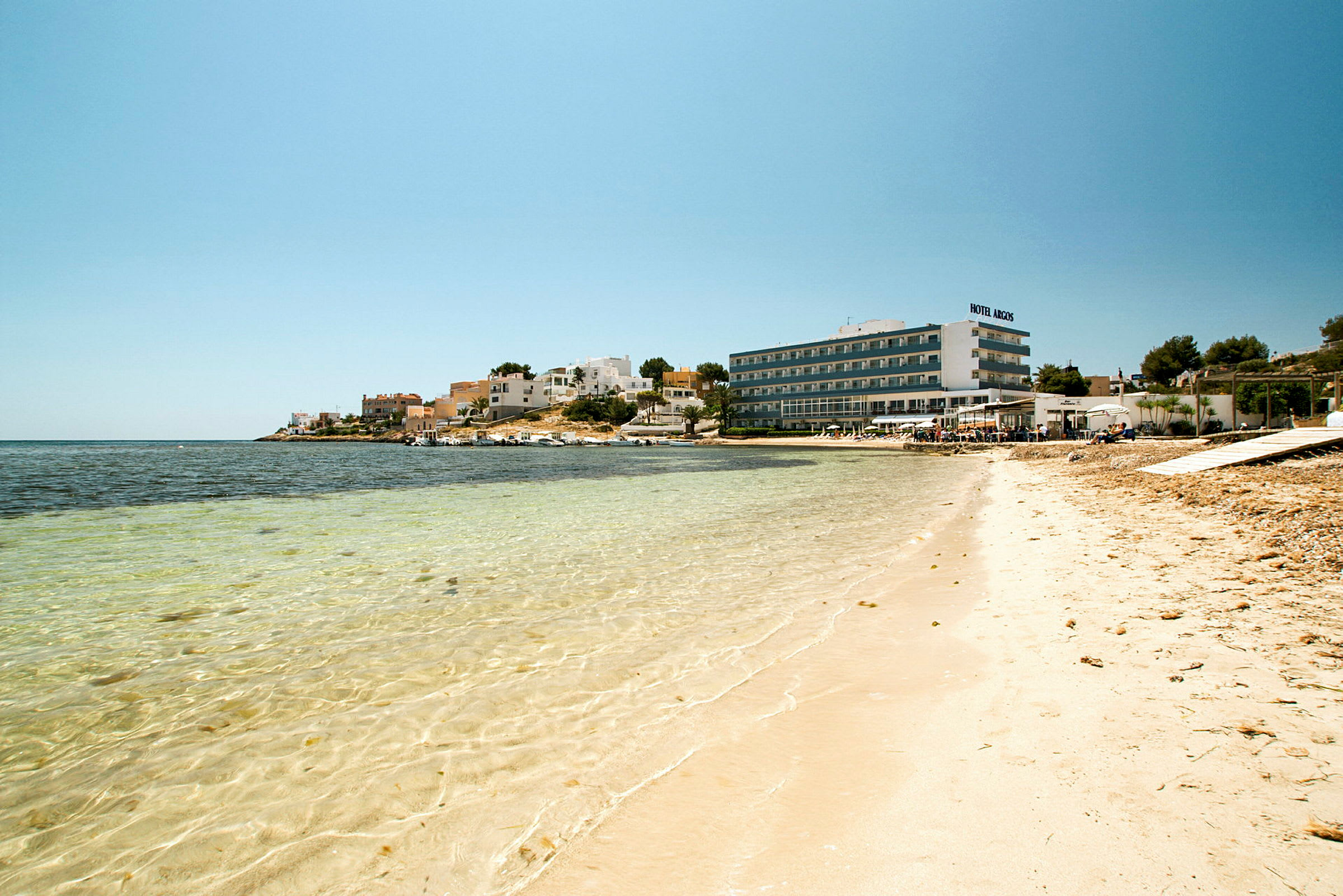 En strand med klart vand, bygninger og et hotel i baggrunden. (AI-genereret tekst)