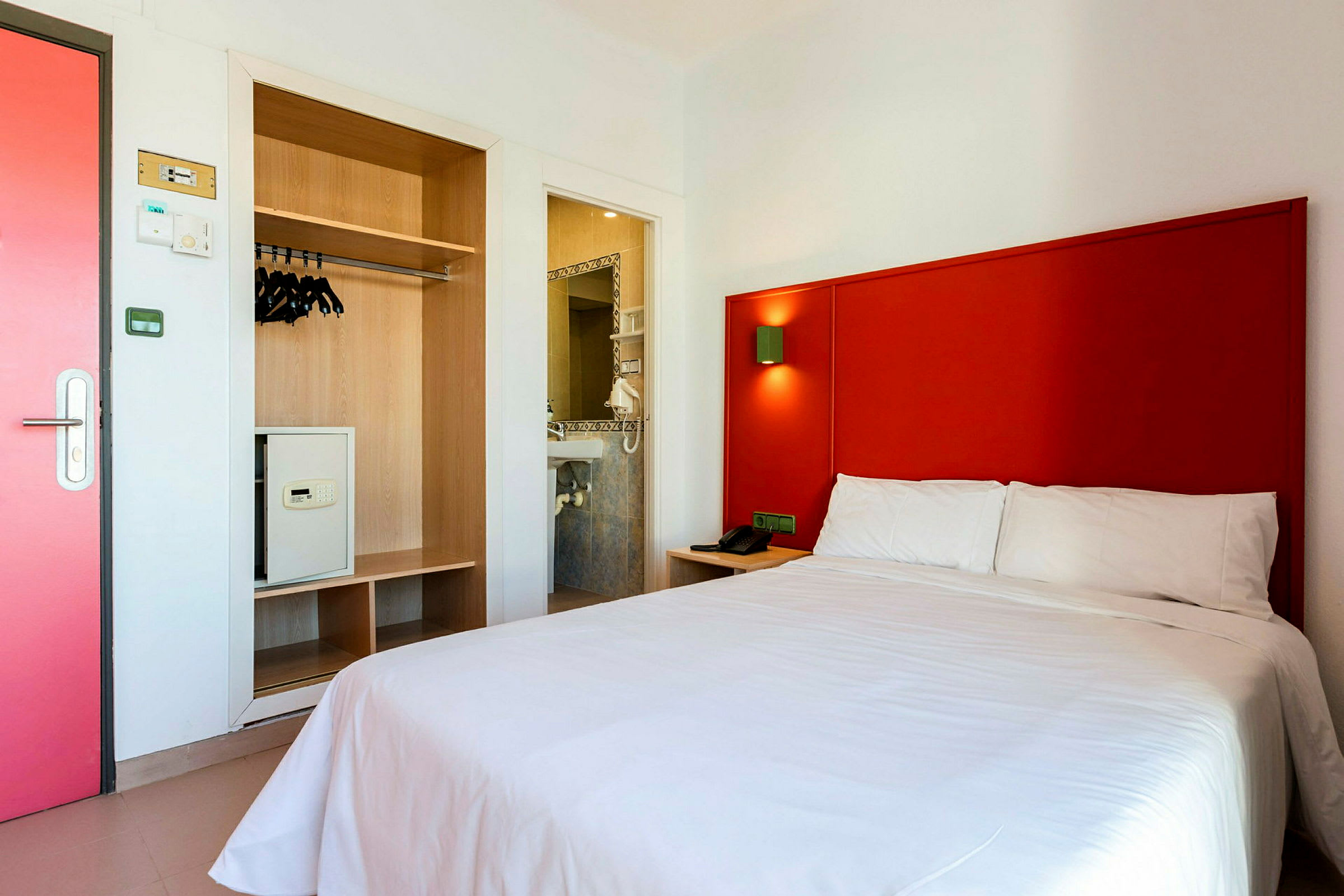 Hotelværelse med rød sengegavl, hvid seng og garderobe med sikkerhedsboks. (AI-genereret tekst)