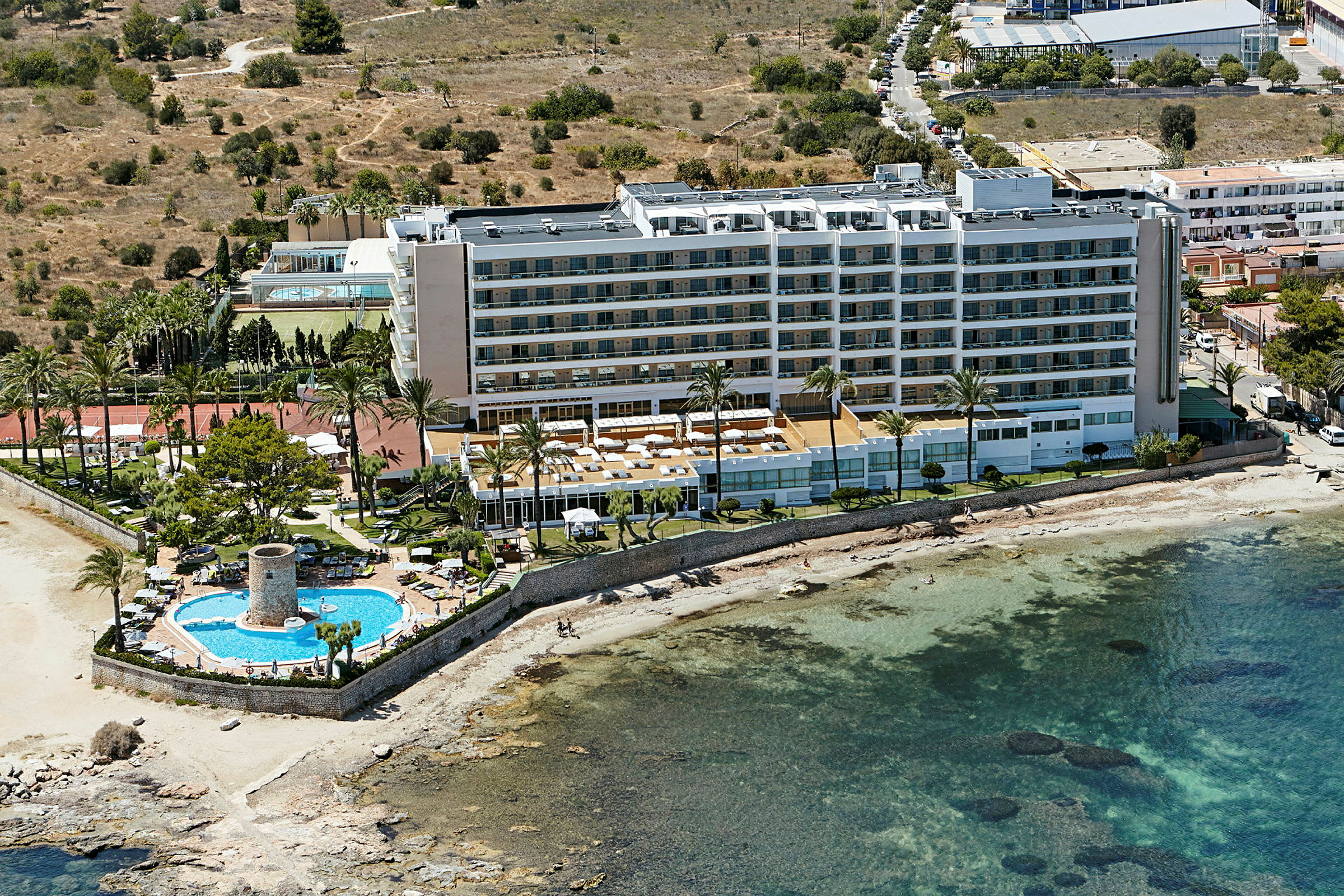 Et hotel ved havet med en pool og liggestole samt omkringliggende grønne områder. (AI-genereret tekst)