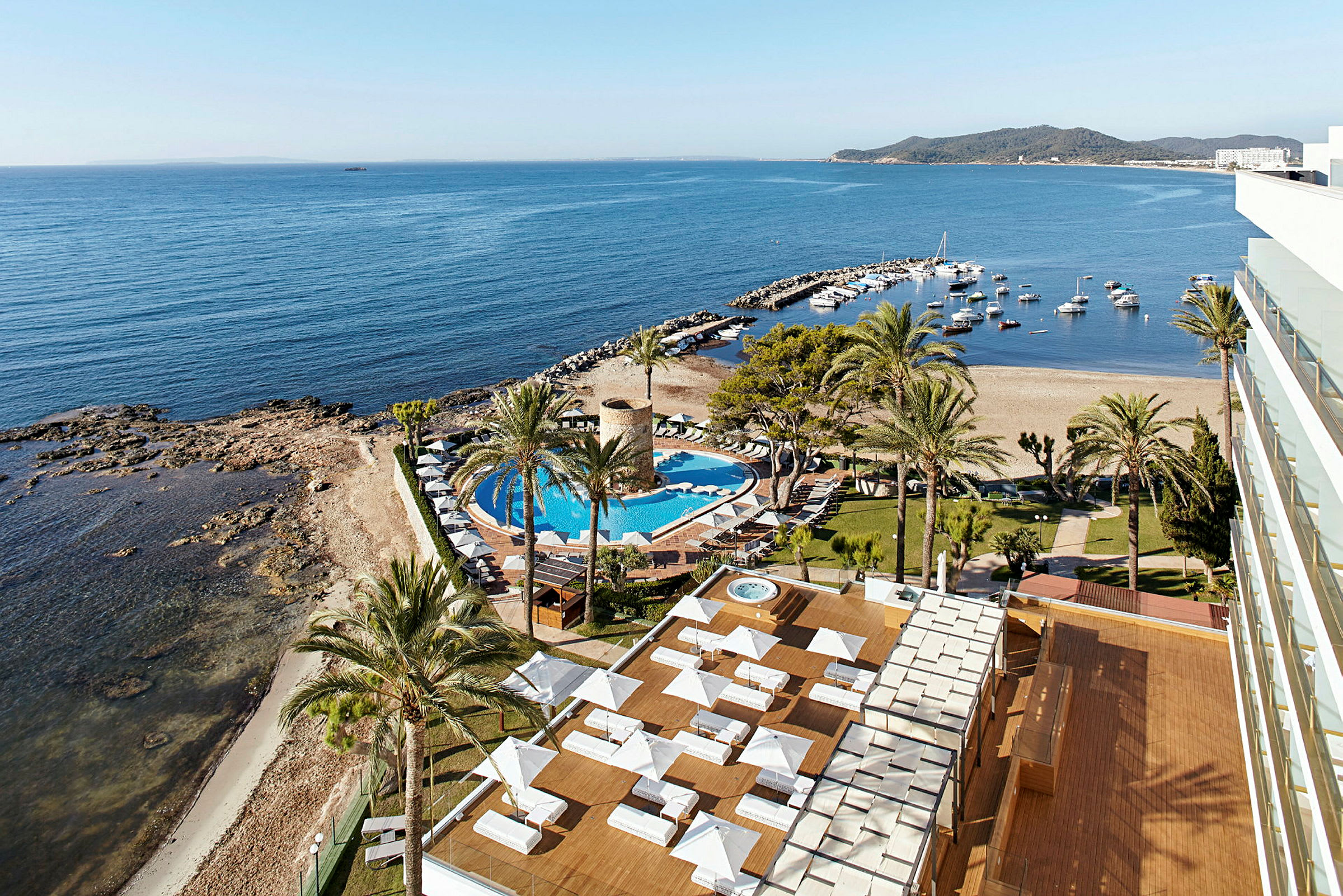 Billedets motiv viser en udsigt over et hotelområde ved havet med en pool, liggestole samt en strand og lystbådehavn i baggrunden. (AI-genereret tekst)