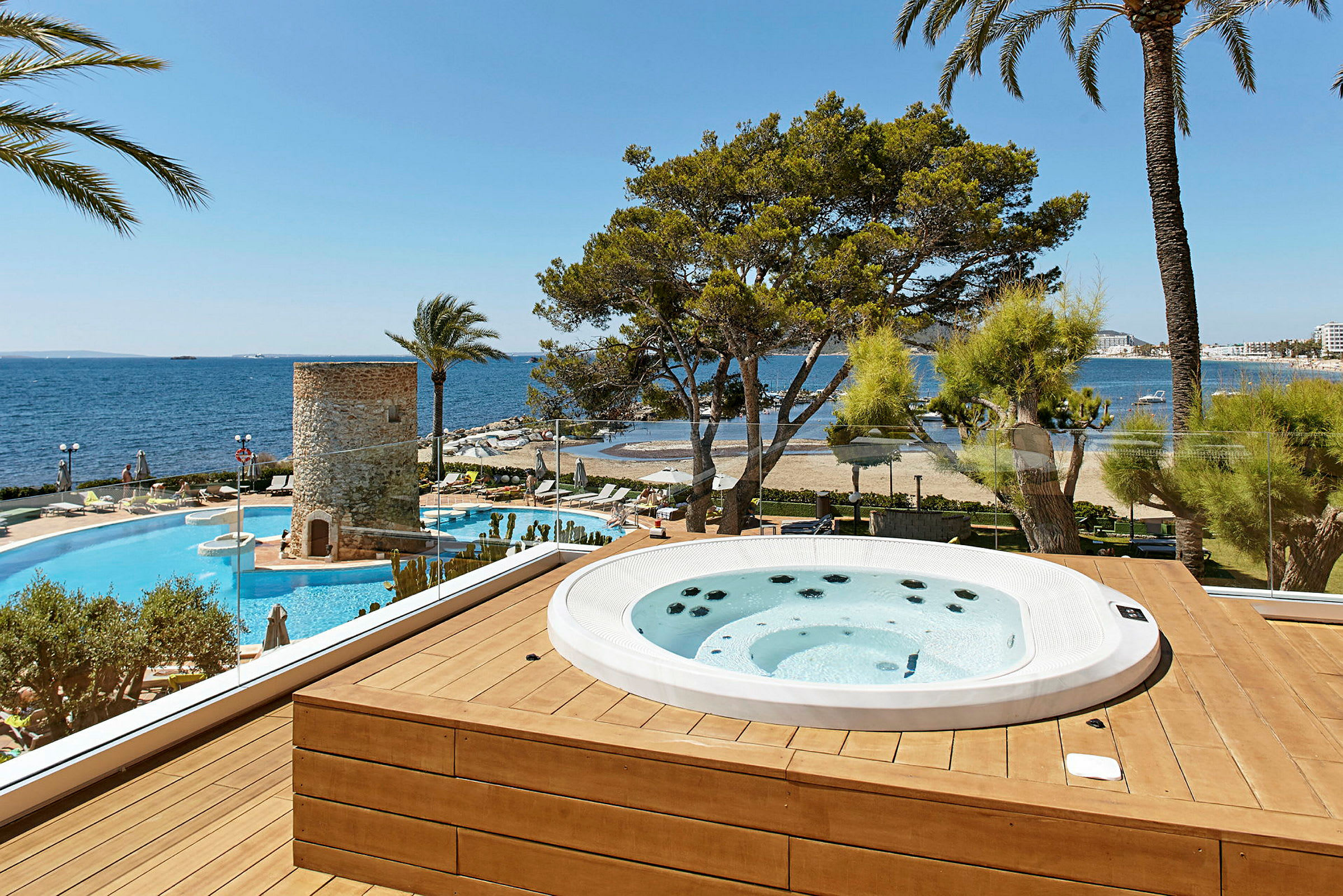 Et spabad på en træterrasse med udsigt over en pool, en strand og havet. Der er også palmer og træer. (AI-genereret tekst)