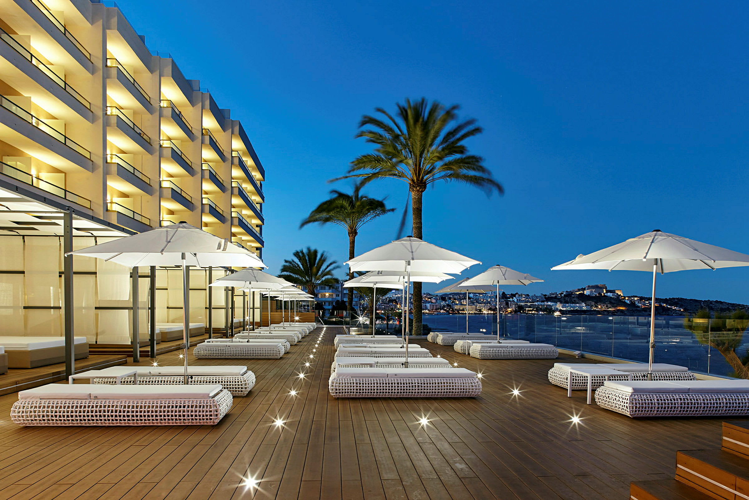 En hotelterrasse om aftenen med udendørs sofaer, hvide parasoller og udsigt over havet og byen. (AI-genereret tekst)