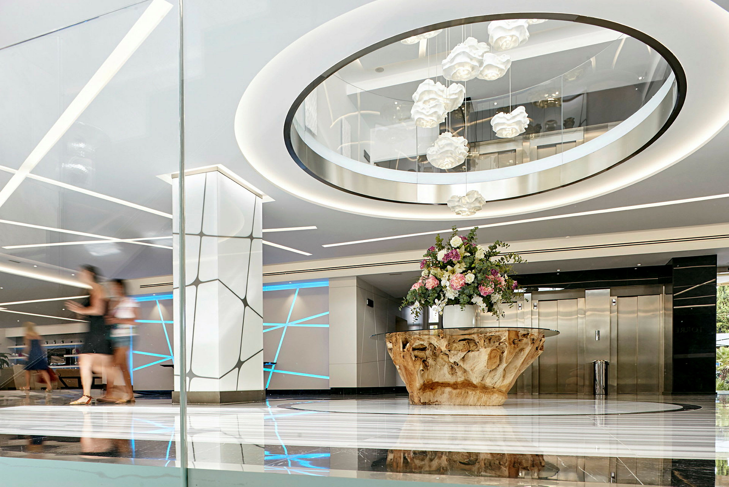 Lys og moderne hotellobby med et stort bord af træ og en buket blomster i midten, elegante lyseffekter og elevatorer i baggrunden. (AI-genereret tekst)