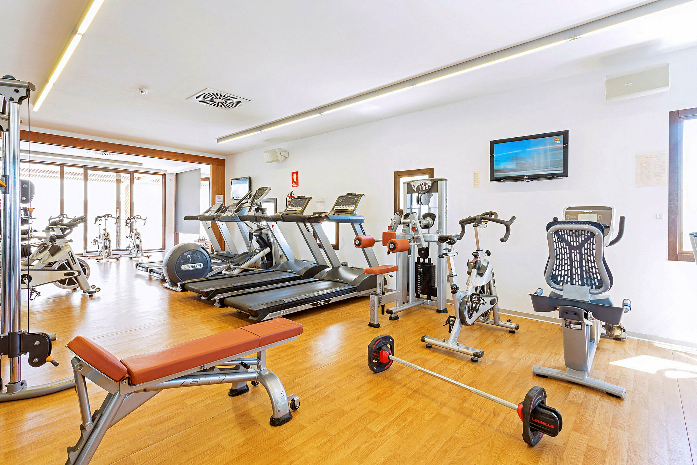 Et fitnesscenter med løbebånd, motionscykler og træningsmaskiner på et lyst trægulv. (AI-genereret tekst)