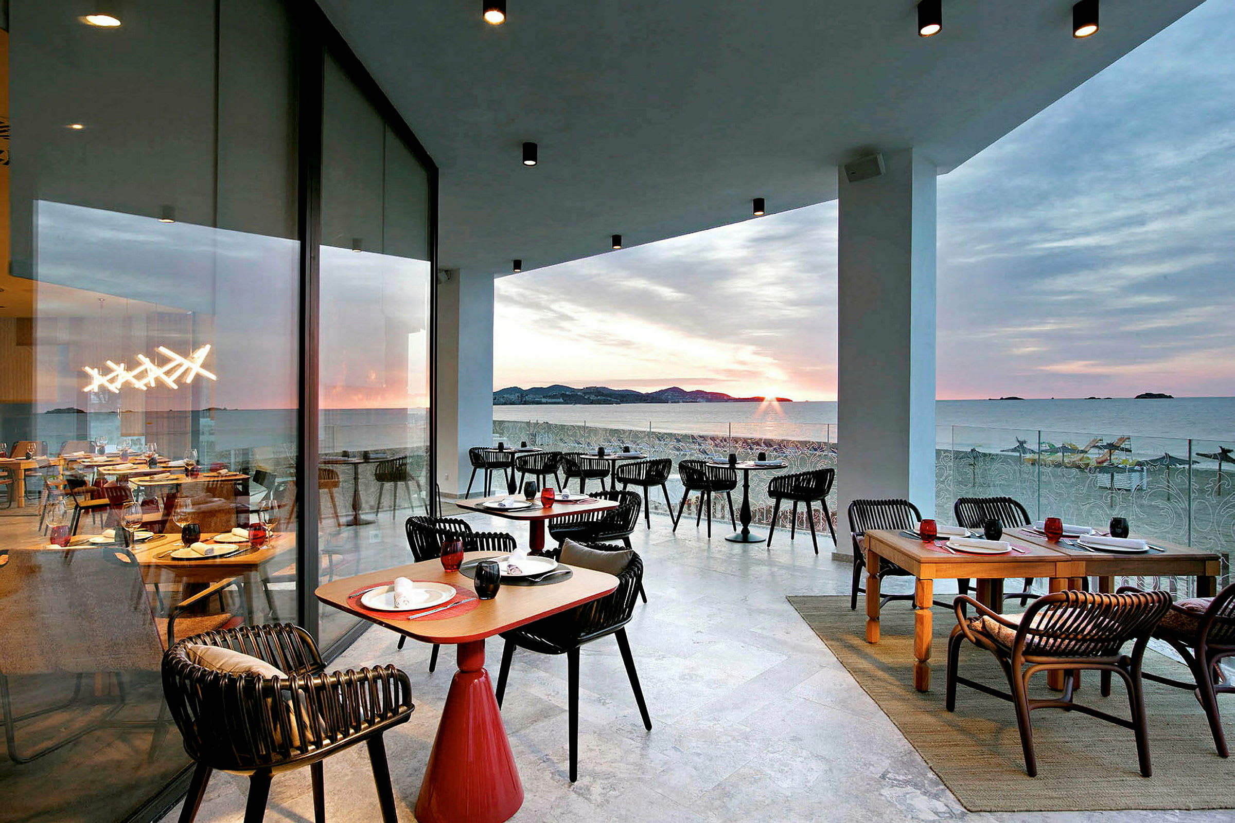 En terrasse ved havet med siddepladser og borde, solnedgang i baggrunden. (AI-genereret tekst)