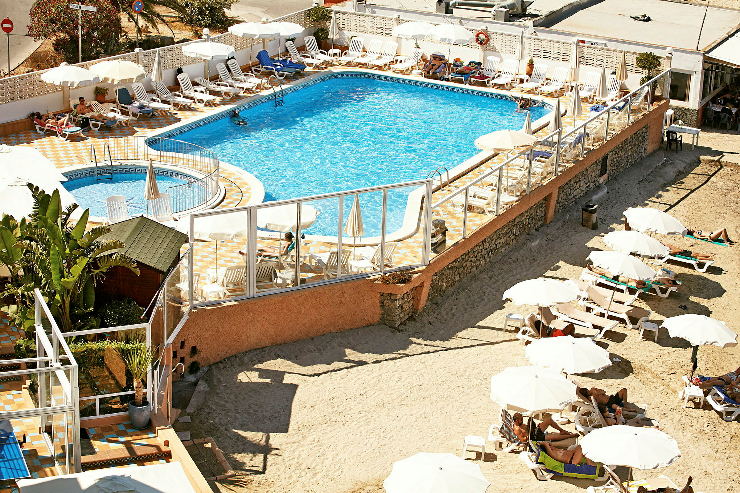 En pool omgivet af liggestole og parasoller, med et sandet område og flere liggestole udenfor området. (AI-genereret tekst)