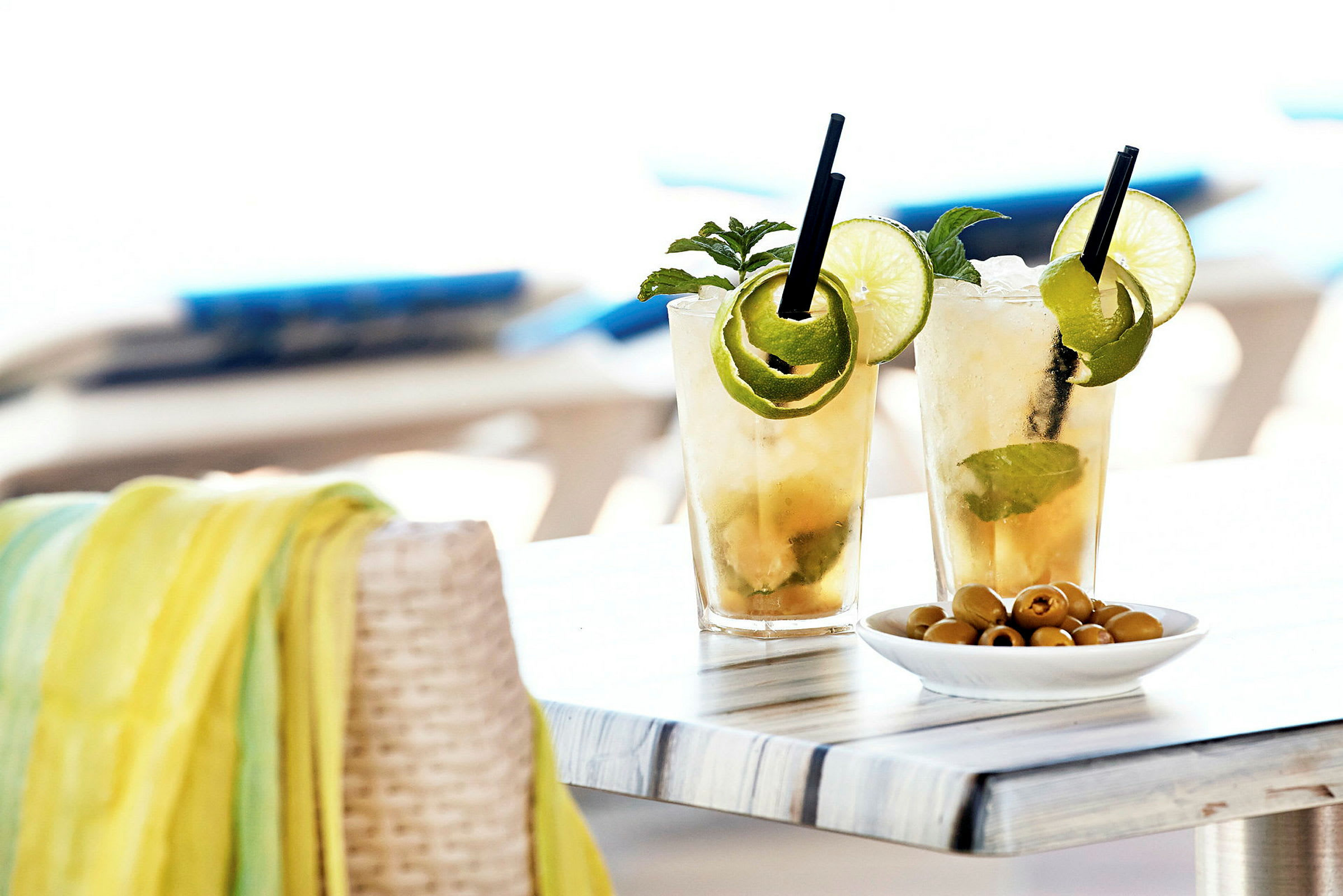 To glas med kolde cocktails pyntet med lime og mynte står på et bord ved siden af en skål med oliven. (AI-genereret tekst)