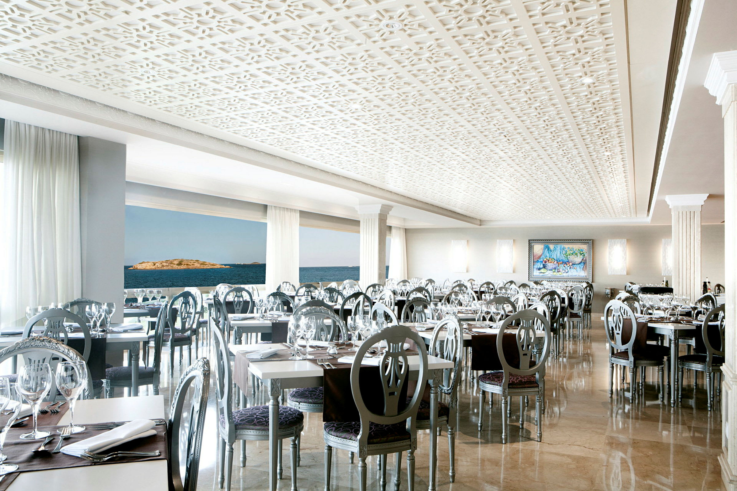 En elegant indrettet restaurant med moderne stole og hvide borde, med store vinduer, der viser udsigten over havet. (AI-genereret tekst)