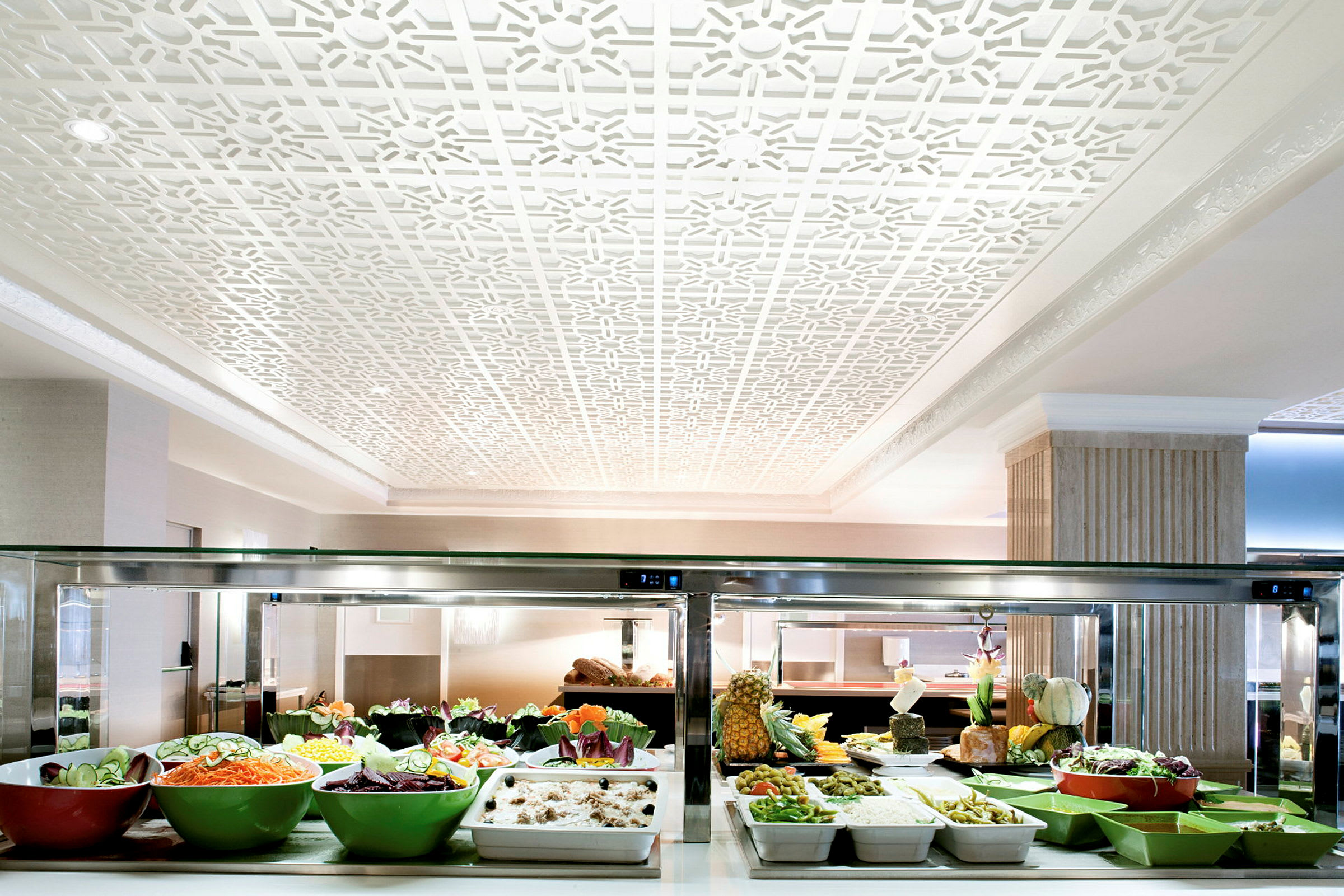 En buffet med forskellige salater og retter i skåle og beholdere, omgivet af et elegant indrettet rum med hvid dekoration i loftet. (AI-genereret tekst)