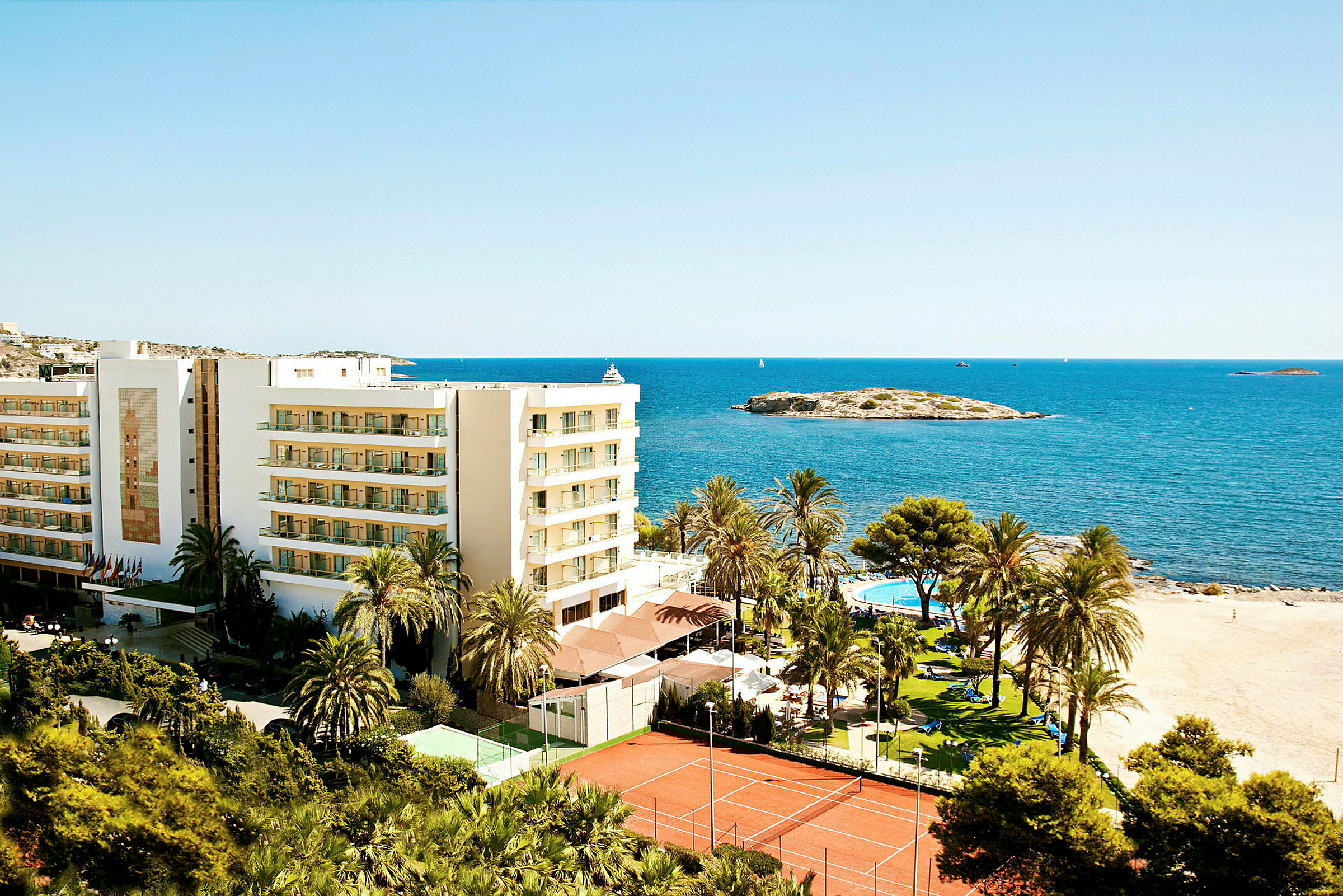 Billede af et feriested ved stranden med en bygning, palmer, swimmingpool og tennisbaner, placeret ved havet. (AI-genereret tekst)
