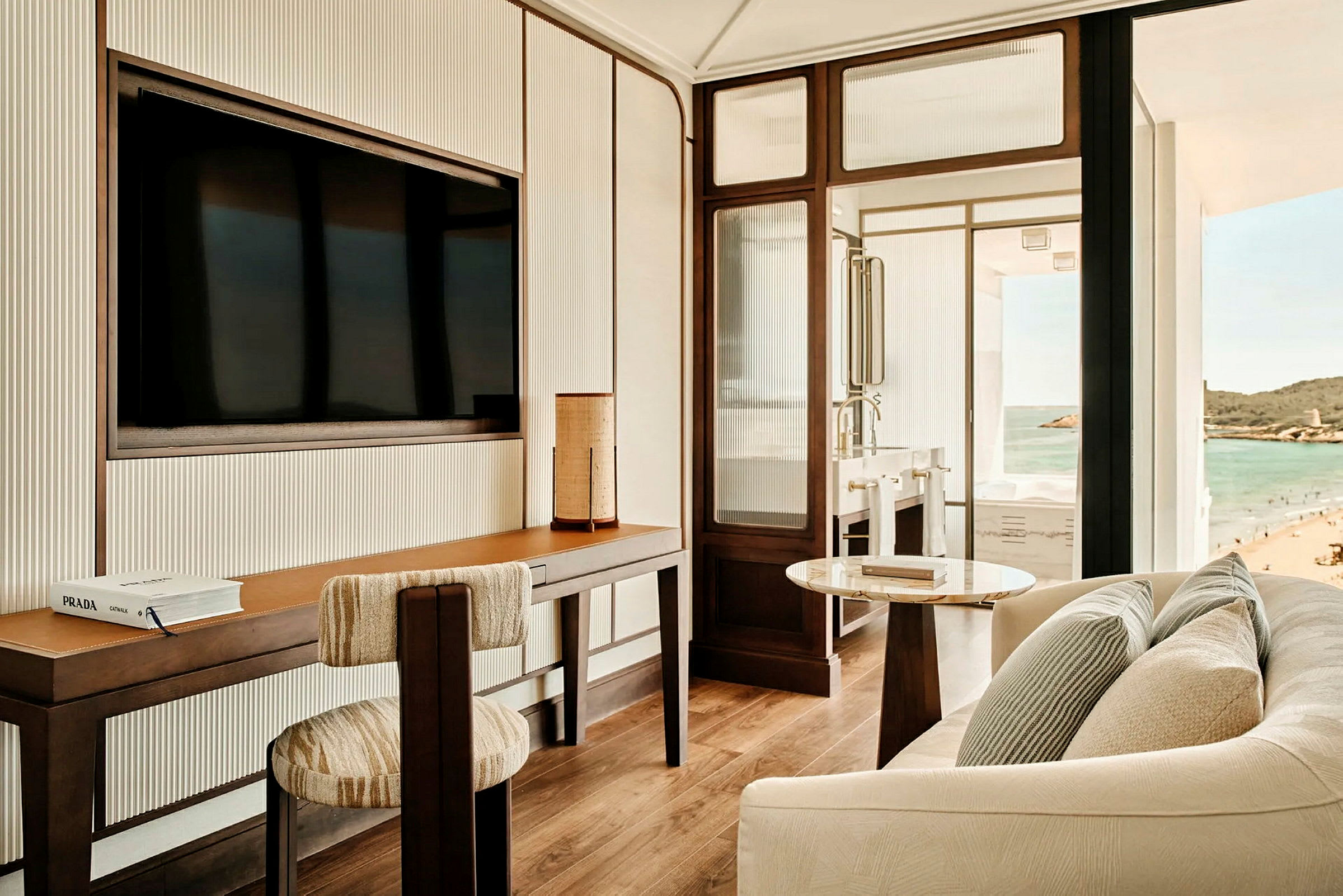 Et moderne hotelværelse med skrivebord, stol, sofa og udsigt over stranden og havet fra vinduet. (AI-genereret tekst)