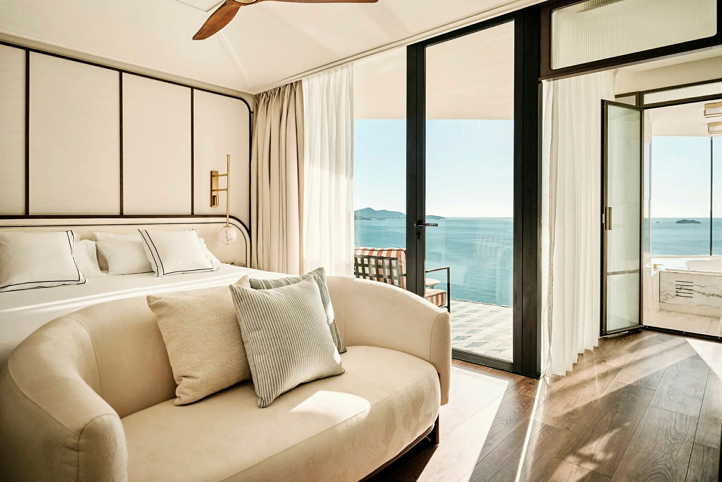 Moderne indrettet hotelværelse med stor seng, sofa og udsigt over havet fra en altan. (AI-genereret tekst)